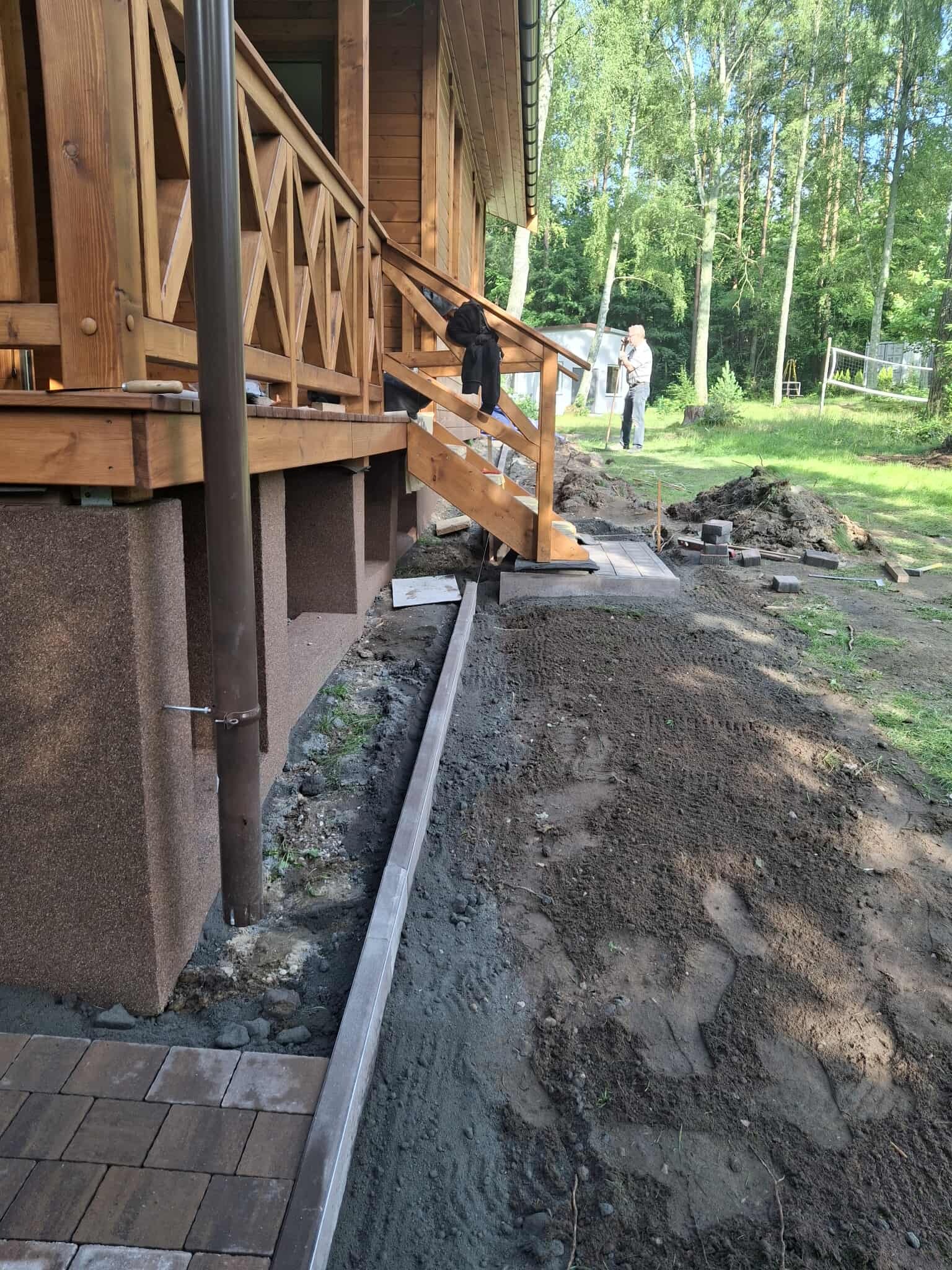 Fragment układania kostki brukowej przy drewnianym domu z werandą. Widoczne obrzeża i przygotowany teren. W tle osoby i zieleń.