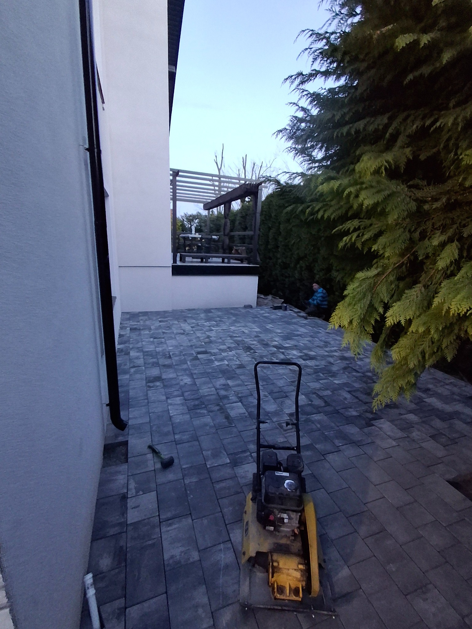 Ułożona kostka brukowa w szarym odcieniu, z ubijarką w tle, obok budynku z pergolą i zielenią. Widoczny fragment pracującego brukarza.