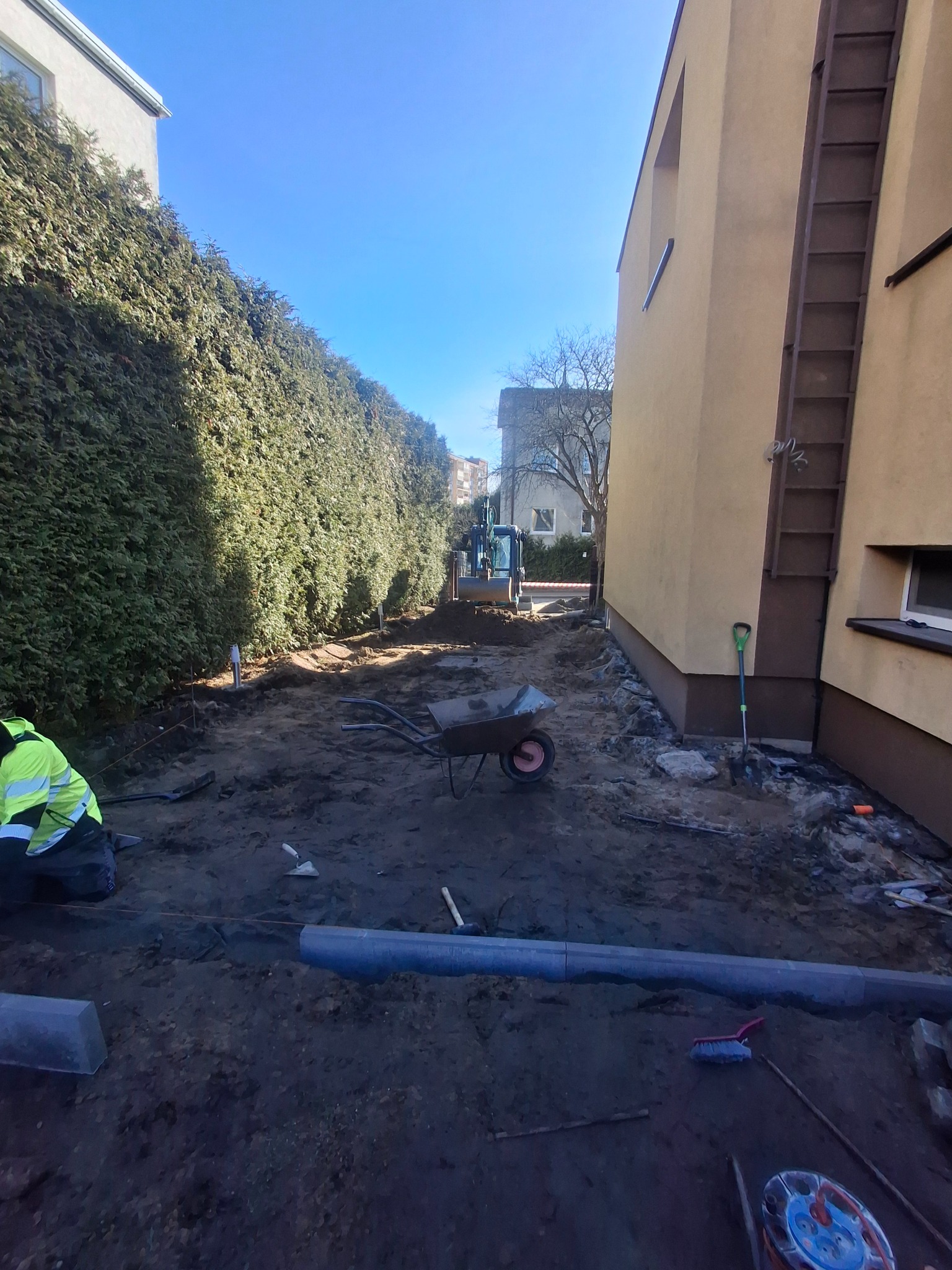 Przygotowanie terenu pod kostkę brukową: wykop, obrzeża, narzędzia, w tle koparka. Widok z perspektywy pracownika w odblaskowej kurtce, wzdłuż żywopłotu i budynku.