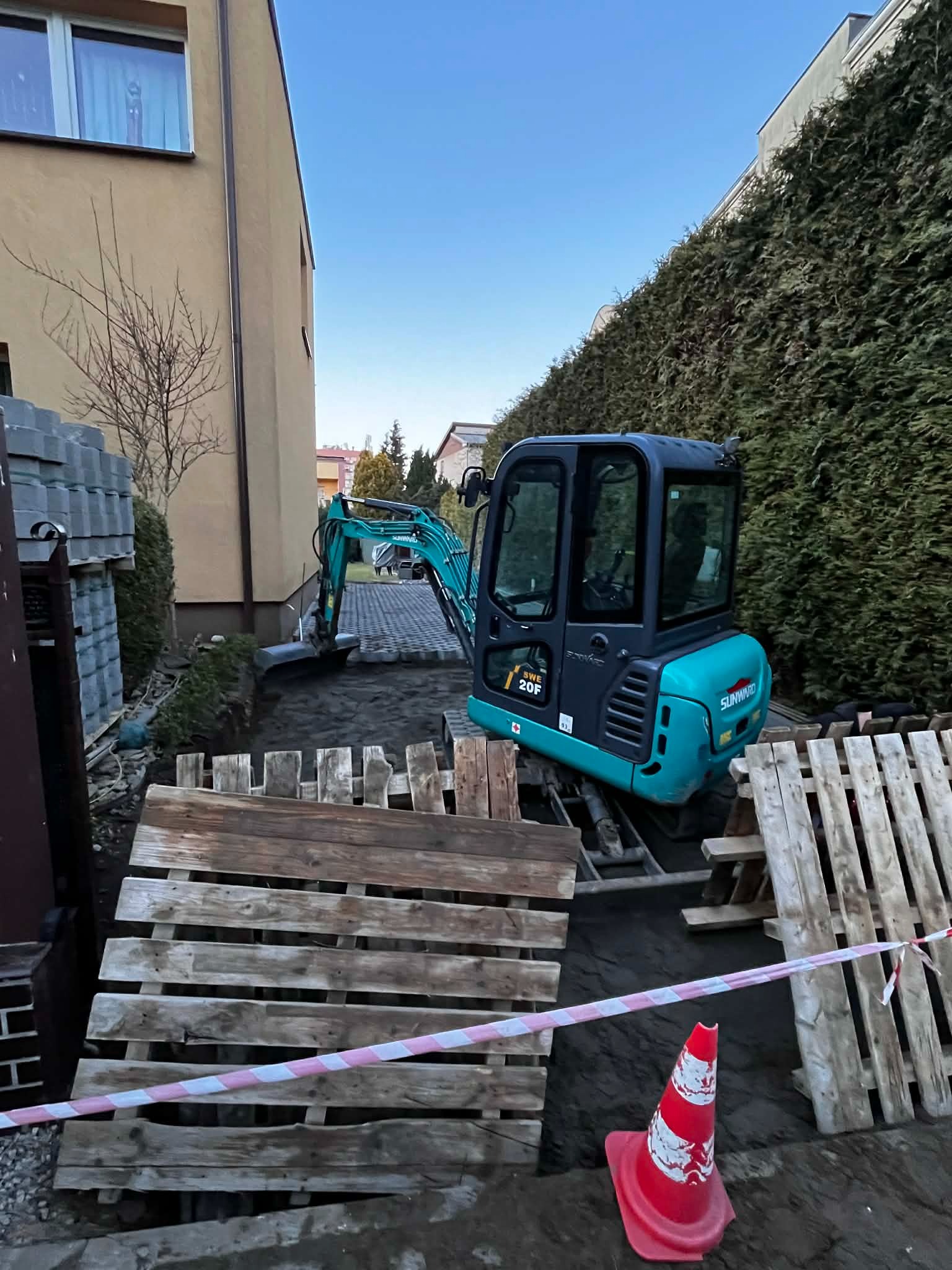 Niebieska mini koparka Sunward SWE 20F na paletach, przygotowująca teren pod budowę, z widocznymi kostkami brukowymi w tle i czerwoną barierką.