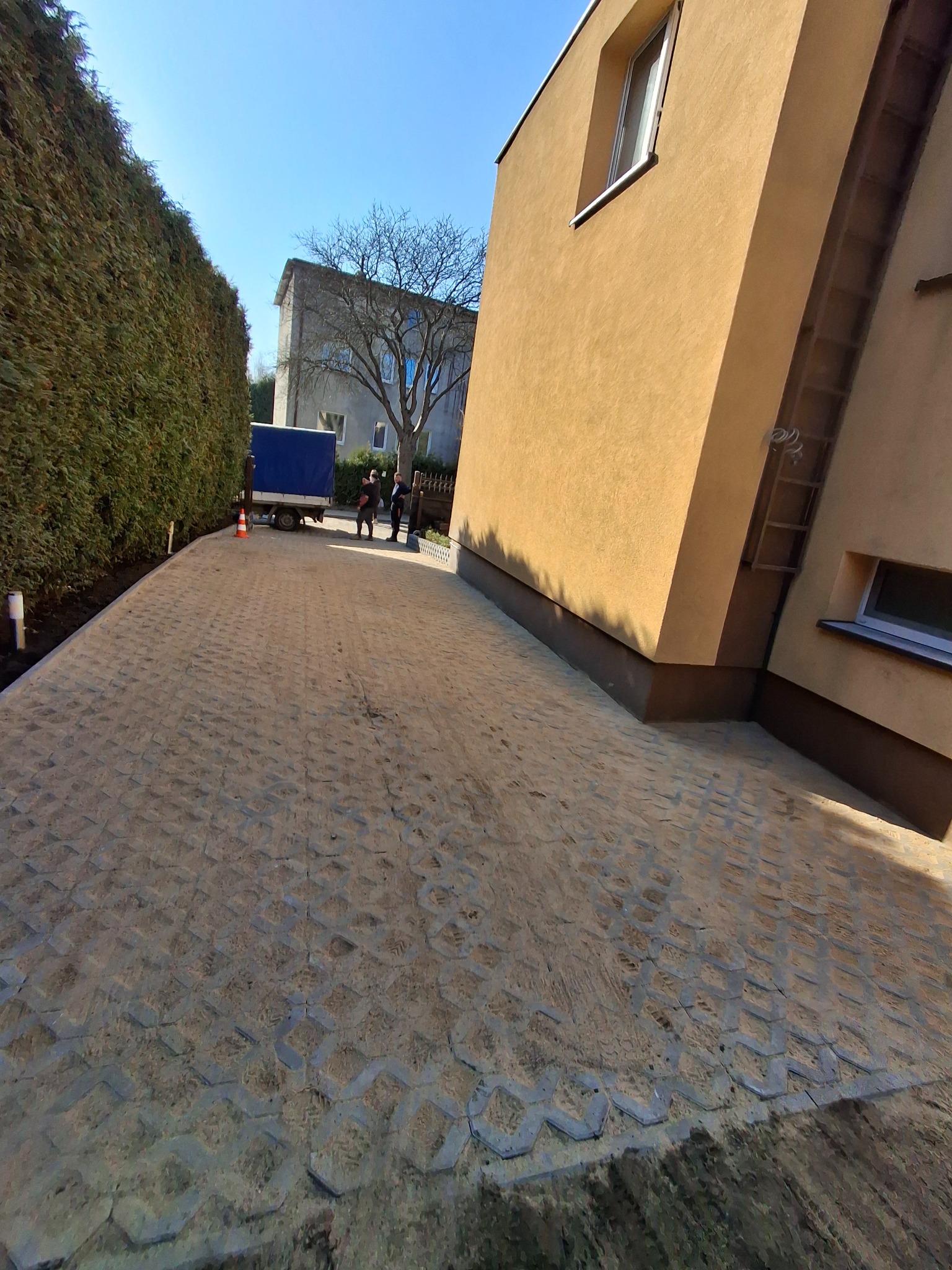 Ułożona, przepuszczalna kostka brukowa na podjeździe, z widocznym budynkiem i zielonym żywopłotem w tle. Na końcu podjazdu stoi przyczepa.