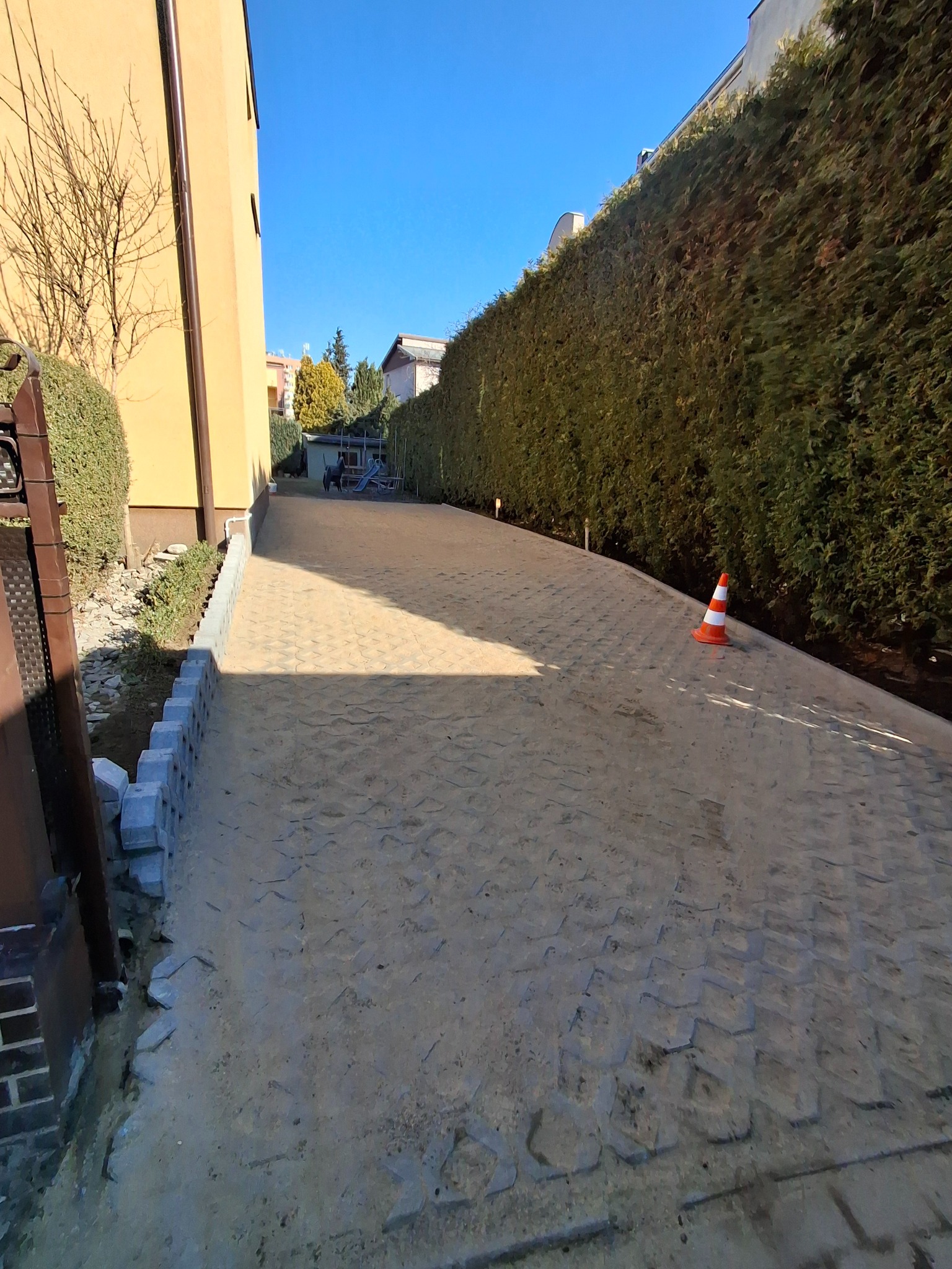 Układanie kostki brukowej na podjeździe, widoczny etap prac z częściowo ułożonym obrzeżem i kostką. Na końcu podjazdu widoczny plac zabaw.