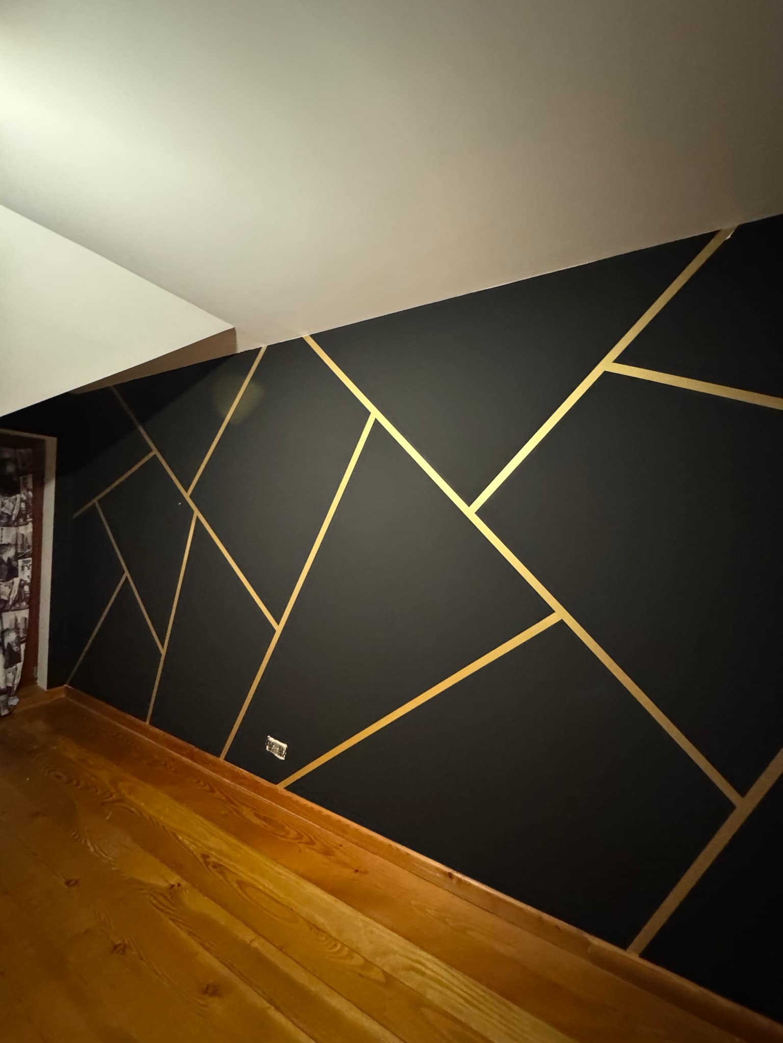 Czarna ściana z geometrycznym wzorem z złotych linii, drewniana podłoga, widok z dołu, gniazdko elektryczne na ścianie.