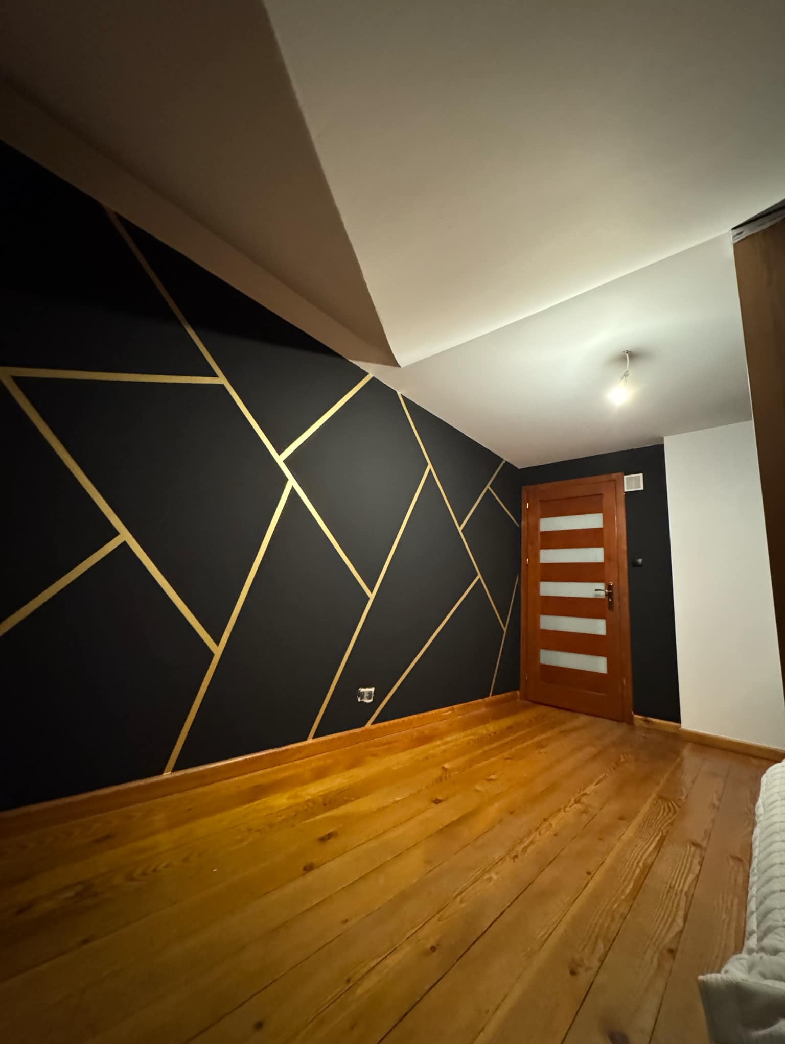 Pokój po remoncie: czarna ściana z geometrycznym wzorem w złotym kolorze, drewniana podłoga i drzwi z przeszkleniami. Ukośny sufit dodaje charakteru.