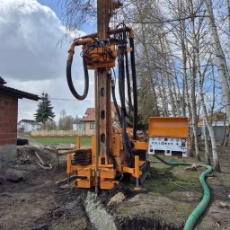 KamComplex Kamil Skrzela - Wiercenie studni głębinowej w Bejsce. Pomarańczowa wiertnica w trakcie pracy, woda wypływa z wykopu. W tle budynek i brzozy.