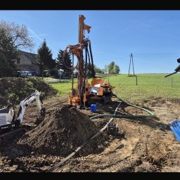 KamComplex Kamil Skrzela - Pomarańczowa wiertnica do studni głębinowych na placu budowy, obok mini koparka Bobcat E10 i niebieskie rury. W tle zielone pole i budynek.