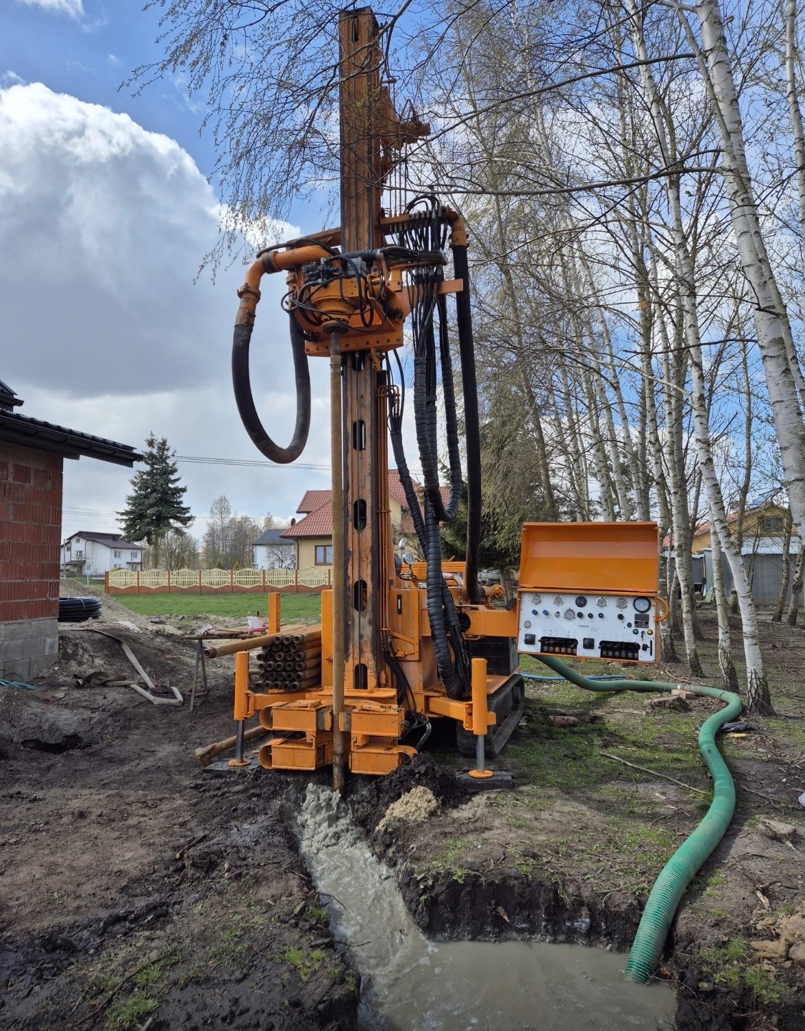 Wiercenie studni głębinowej w Bejsce. Pomarańczowa wiertnica w trakcie pracy, woda wypływa z wykopu. W tle budynek i brzozy.