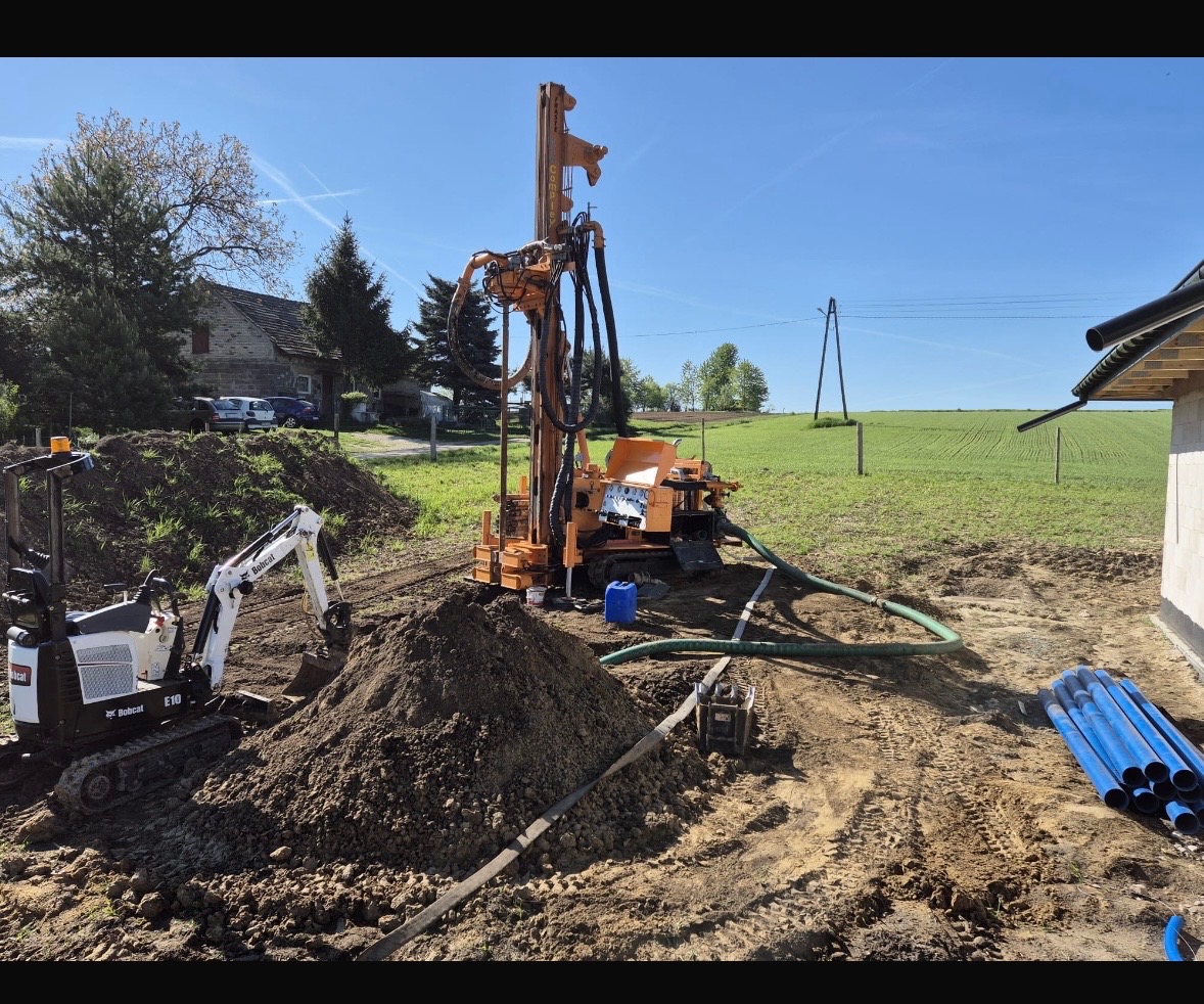 Pomarańczowa wiertnica do studni głębinowych na placu budowy, obok mini koparka Bobcat E10 i niebieskie rury. W tle zielone pole i budynek.