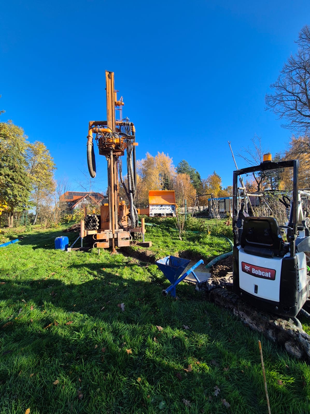 Wiertnica do studni głębinowych na trawniku, obok minikoparka Bobcat. Widoczne rury, błękitne niebo i jesienne drzewa w tle. Prace w toku, teren przygotowany.