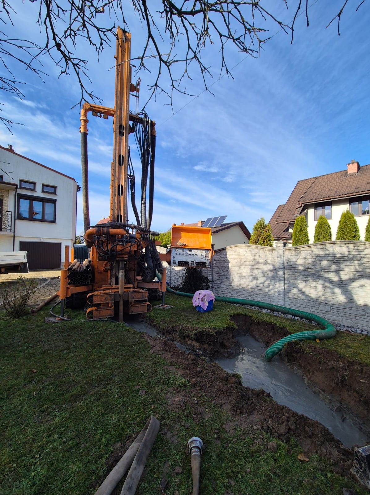 Pomarańczowa wiertnica do studni głębinowych na trawniku, z widocznym wykopem wypełnionym wodą i zielonym wężem odprowadzającym. W tle domy i błękitne niebo.