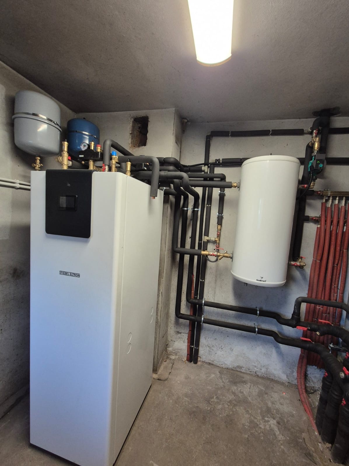 Biała pompa ciepła Stiebel Eltron z zbiornikami i rurami w piwnicy, widoczne elementy instalacji grzewczej i oświetlenie sufitowe.