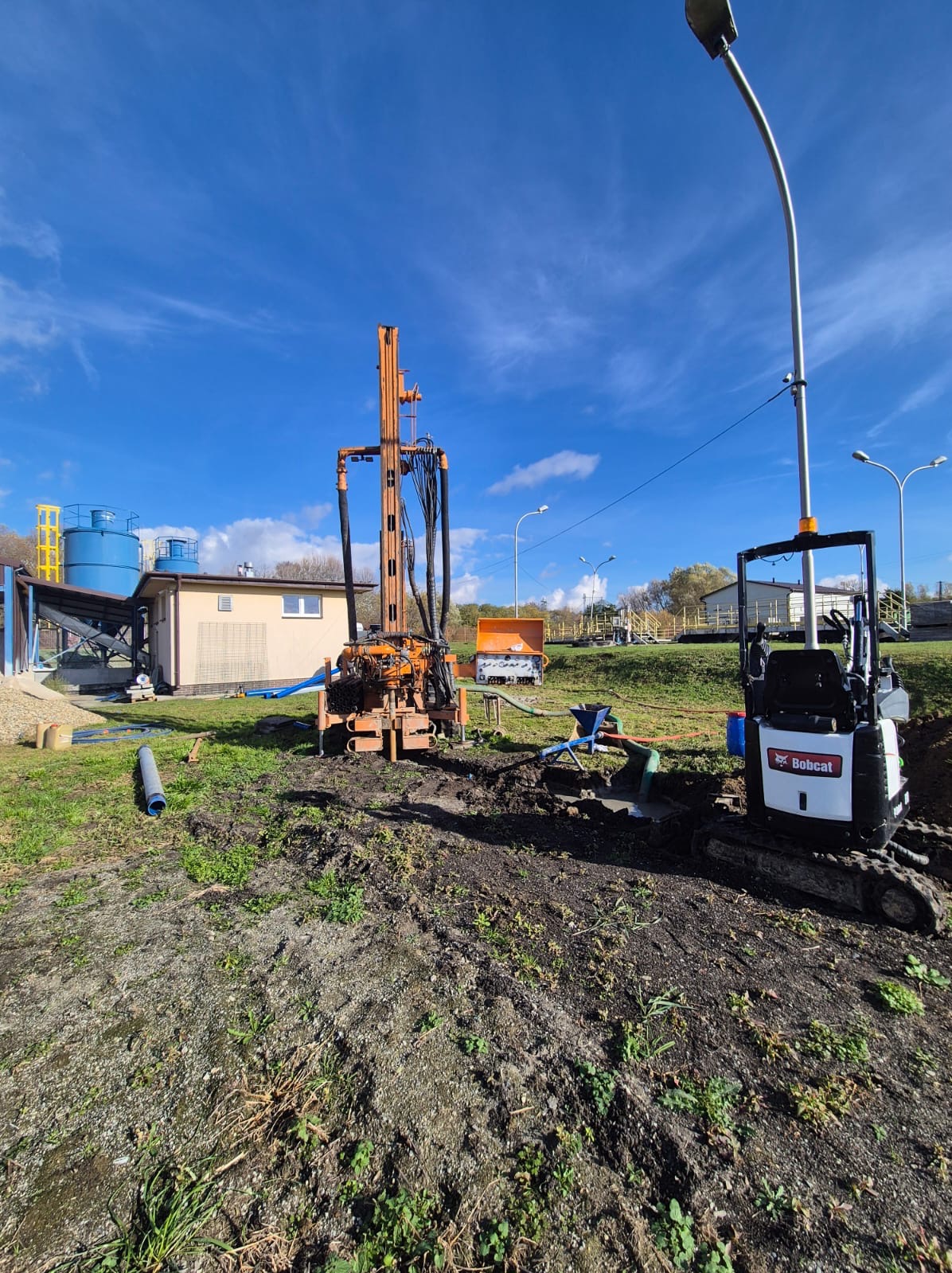 Wiertnica i minikoparka Bobcat na placu budowy w Bejsce. Sprzęt do wiercenia studni głębinowych, błękitne niebo, widoczne elementy infrastruktury.