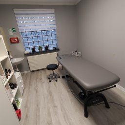 Studio Masażu Wojciech Kabak - Jasne wnętrze gabinetu z szarym łóżkiem do zabiegów, stołkiem na kółkach i oknem z luksferami. Apteczka pierwszej pomocy na ścianie. Minimalistyczny wystrój.