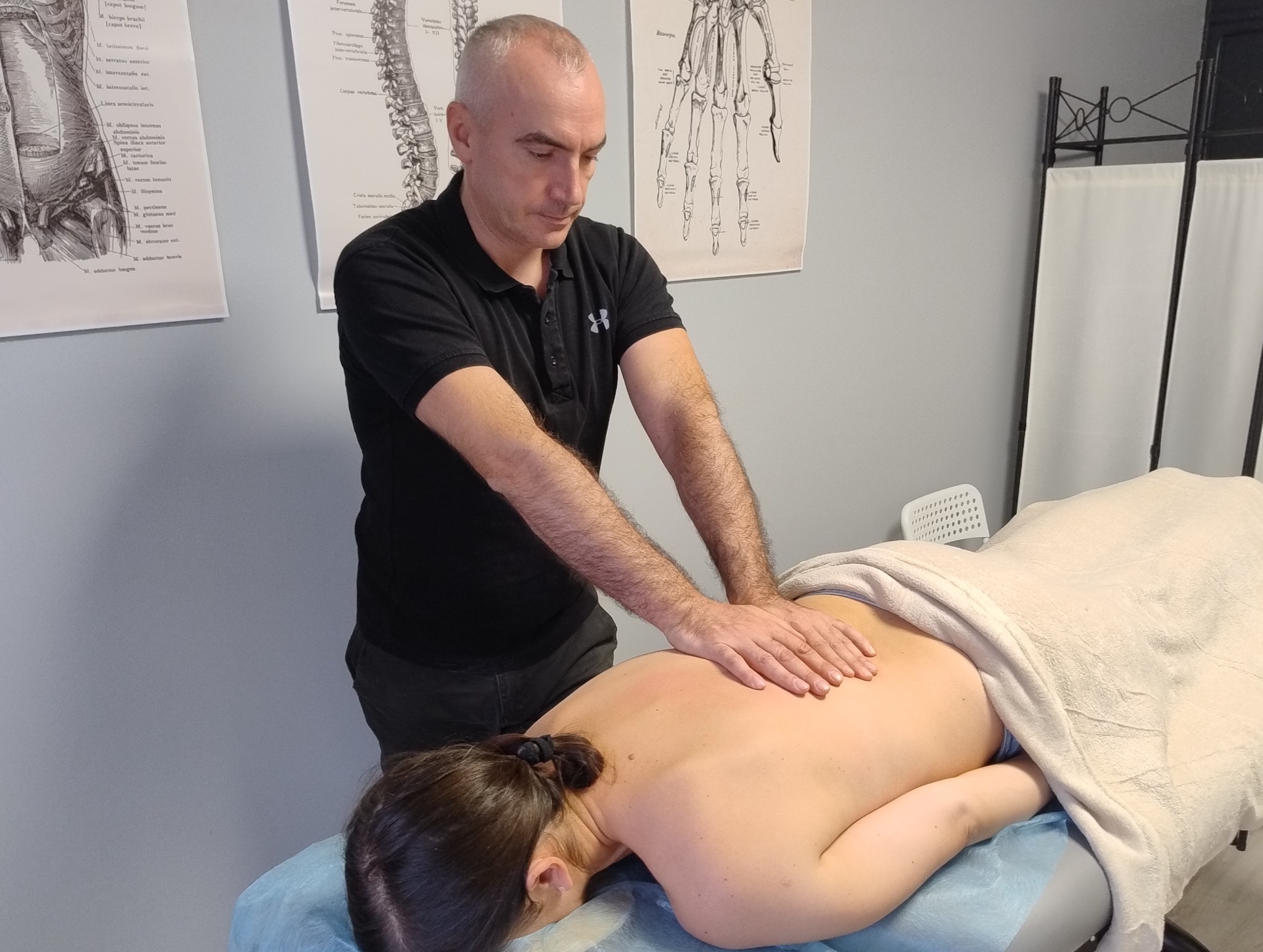 Terapeuta manualny wykonuje masaż pleców pacjentki na kozetce. W tle widoczne plakaty anatomiczne. Skupienie na dłoniach terapeuty i plecach pacjentki.