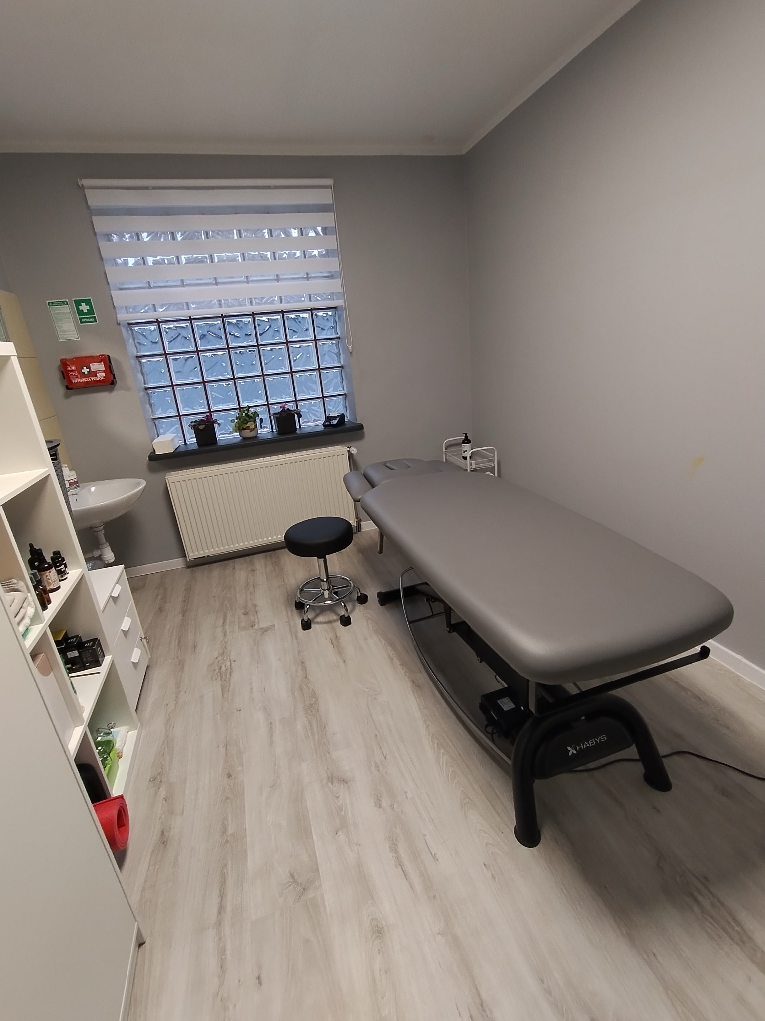 Jasne wnętrze gabinetu z szarym łóżkiem do zabiegów, stołkiem na kółkach i oknem z luksferami. Apteczka pierwszej pomocy na ścianie. Minimalistyczny wystrój.