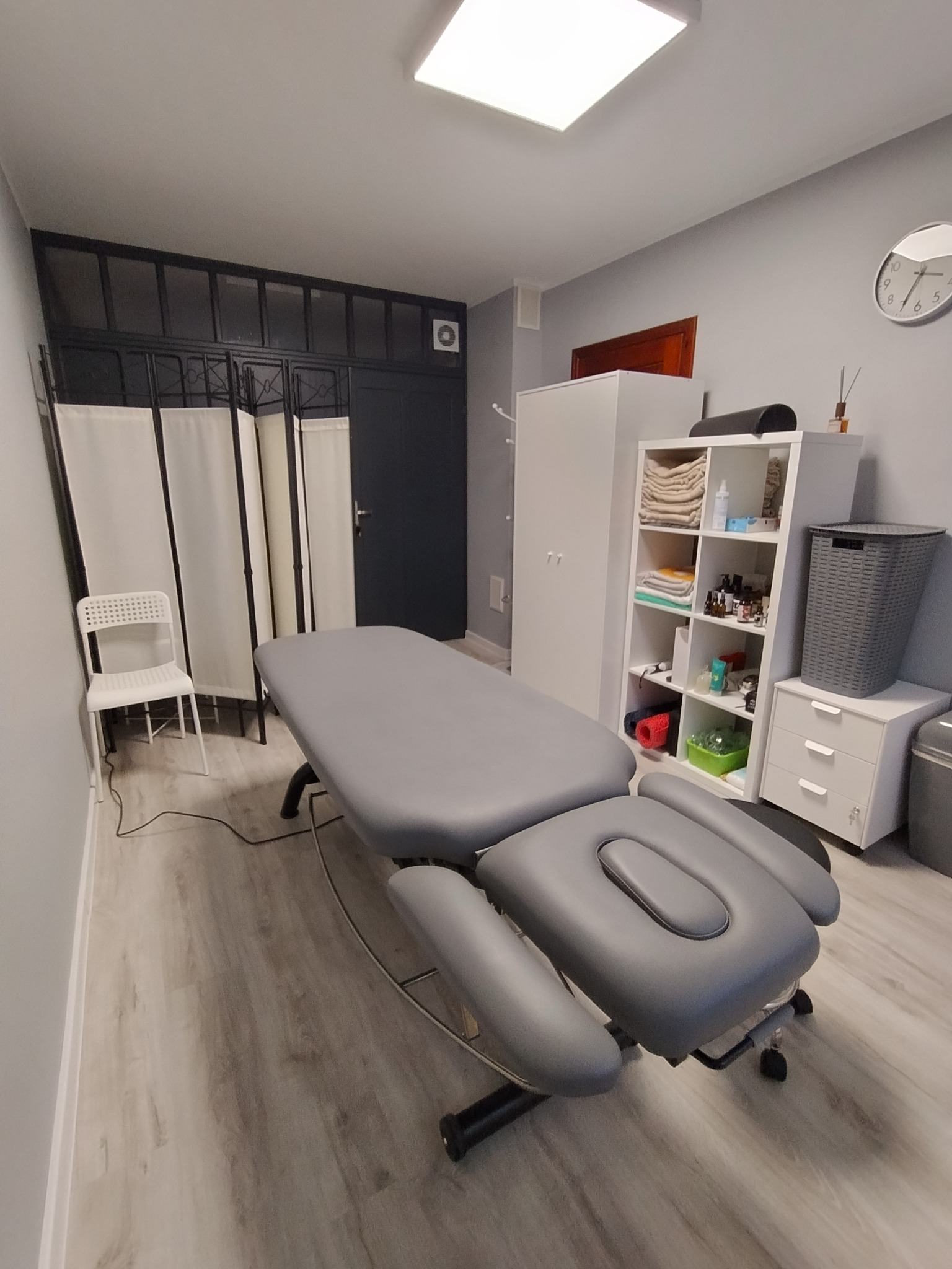 Nowoczesny gabinet z szarym stołem do zabiegów, parawanem i białymi meblami. Jasne wnętrze z drewnianą podłogą i minimalistycznym wystrojem.