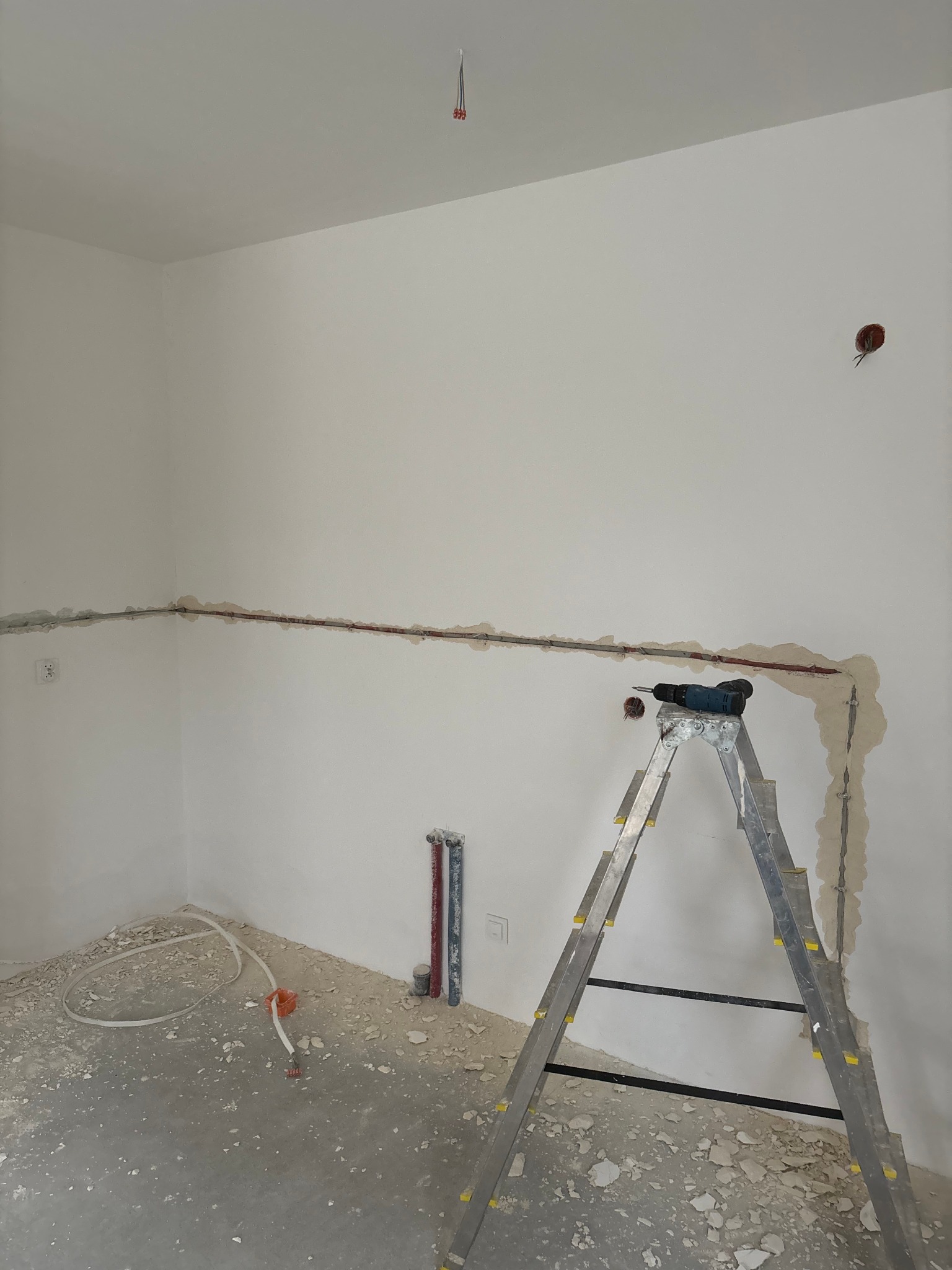 Wnętrze w trakcie prac instalacyjnych: bruzdy w ścianach z przewodami, drabina z wiertarką, widoczne gniazdko i wystające kable z sufitu. Podłoga pokryta gruzem.