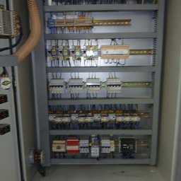 EI-ELEKTRYK Sp. z o.o. - Otwarta szafa sterownicza z widocznymi elementami instalacji elektrycznej, przewodami i zabezpieczeniami. Szara obudowa, liczne kable i moduły.