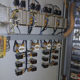 EI-ELEKTRYK Sp. z o.o. - Wnętrze szafy sterowniczej z modułami, przewodami i przełącznikami. Widoczne elementy instalacji elektrycznej w szarej obudowie, z licznymi połączeniami kablowymi.