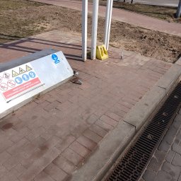 EI-ELEKTRYK Sp. z o.o. - Metalowa obudowa z piktogramami BHP i napisem o odzieży antystatycznej, stojąca na czerwonej kostce brukowej obok kratki ściekowej i białych słupków.