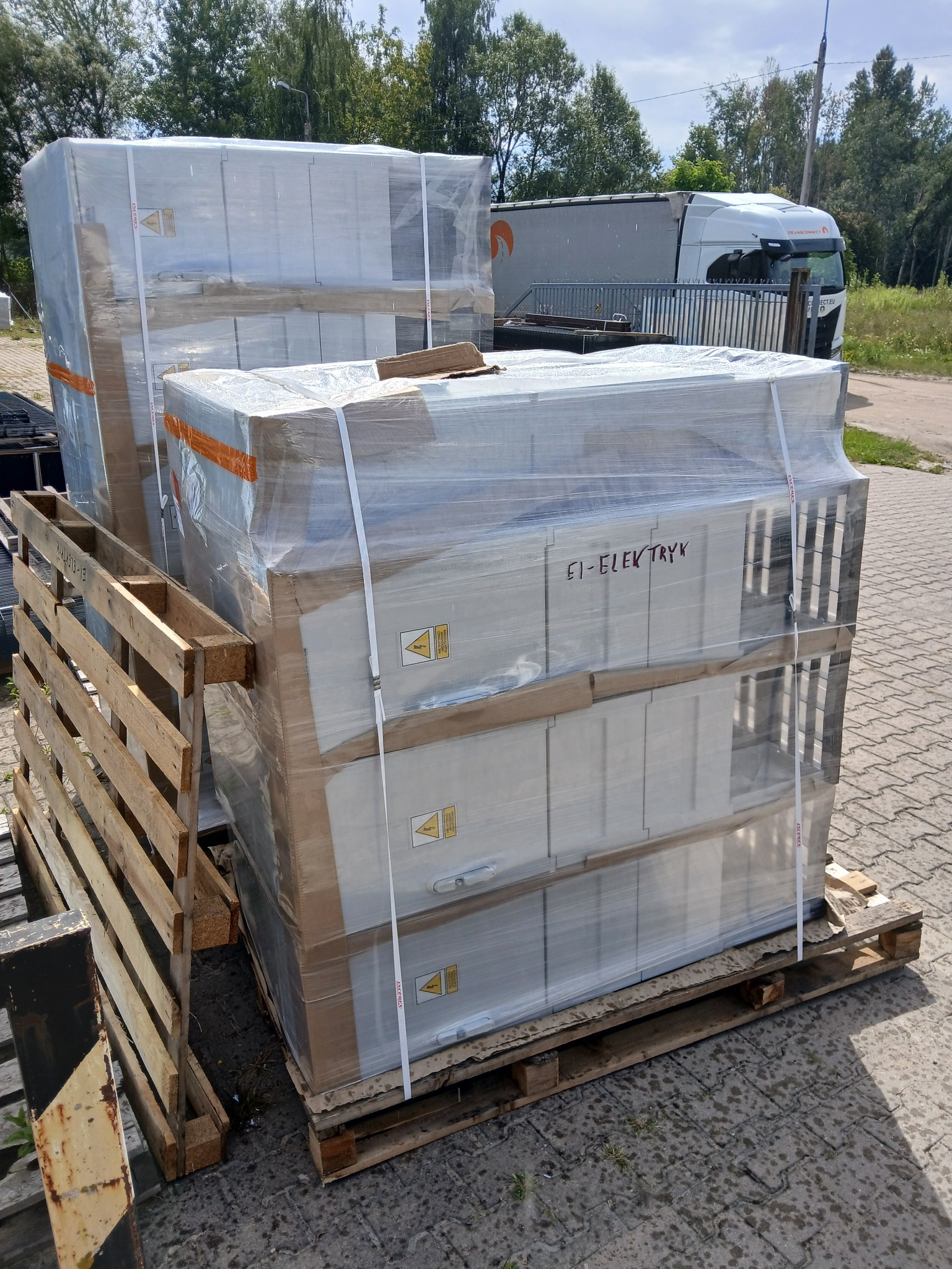 Szafy elektryczne EI-ELEKTRYK, zabezpieczone folią, na paletach. Widoczne znaki ostrzegawcze. W tle ciężarówka i zieleń. Dostawa komponentów instalacji elektrycznych.
