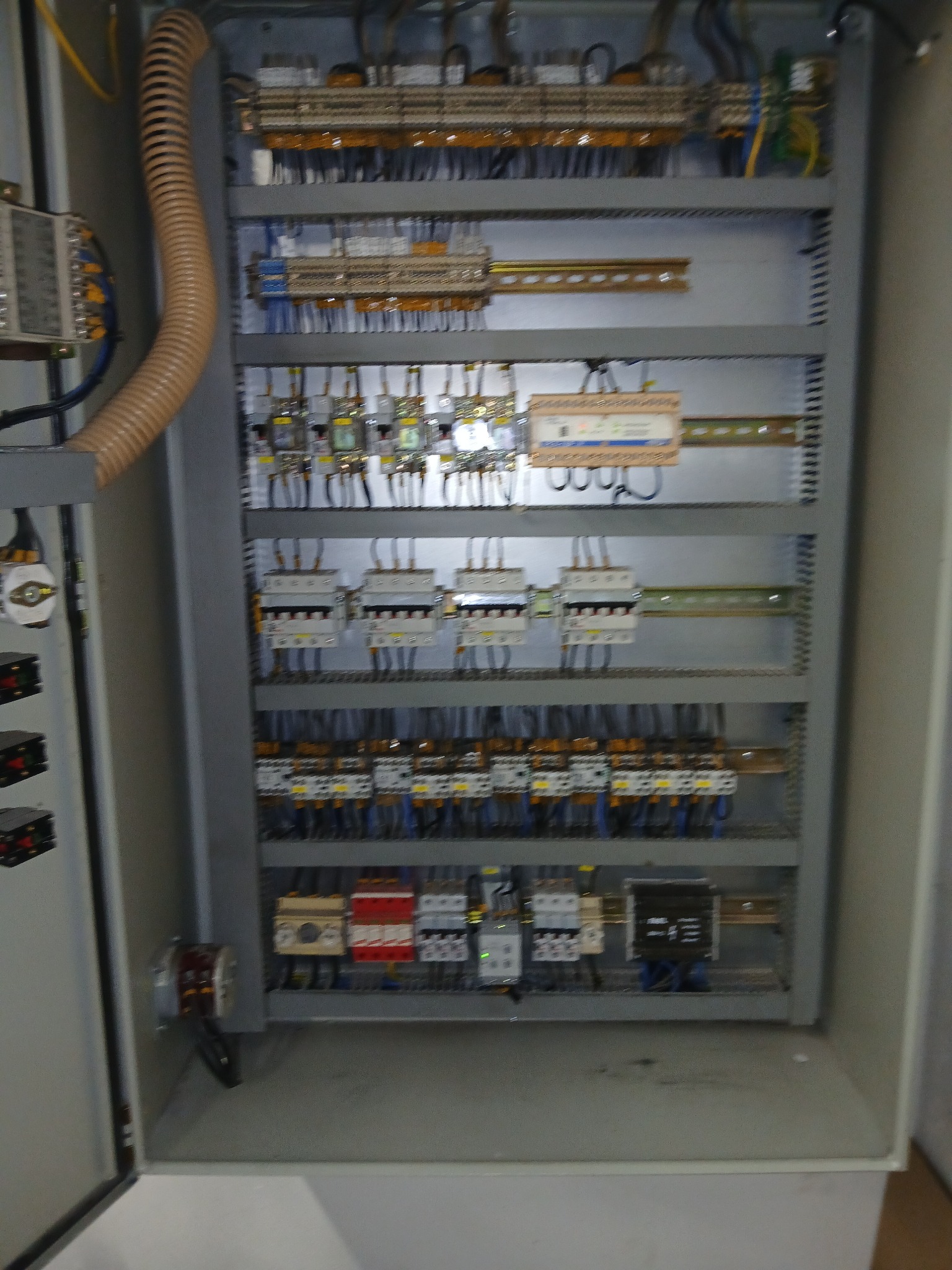 Otwarta szafa sterownicza z widocznymi elementami instalacji elektrycznej, przewodami i zabezpieczeniami. Szara obudowa, liczne kable i moduły.