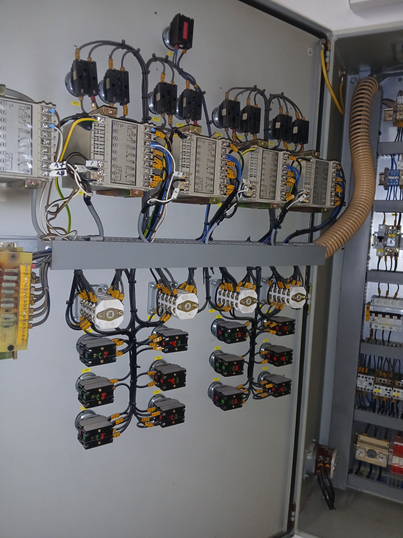 Wnętrze szafy sterowniczej z modułami, przewodami i przełącznikami. Widoczne elementy instalacji elektrycznej w szarej obudowie, z licznymi połączeniami kablowymi.