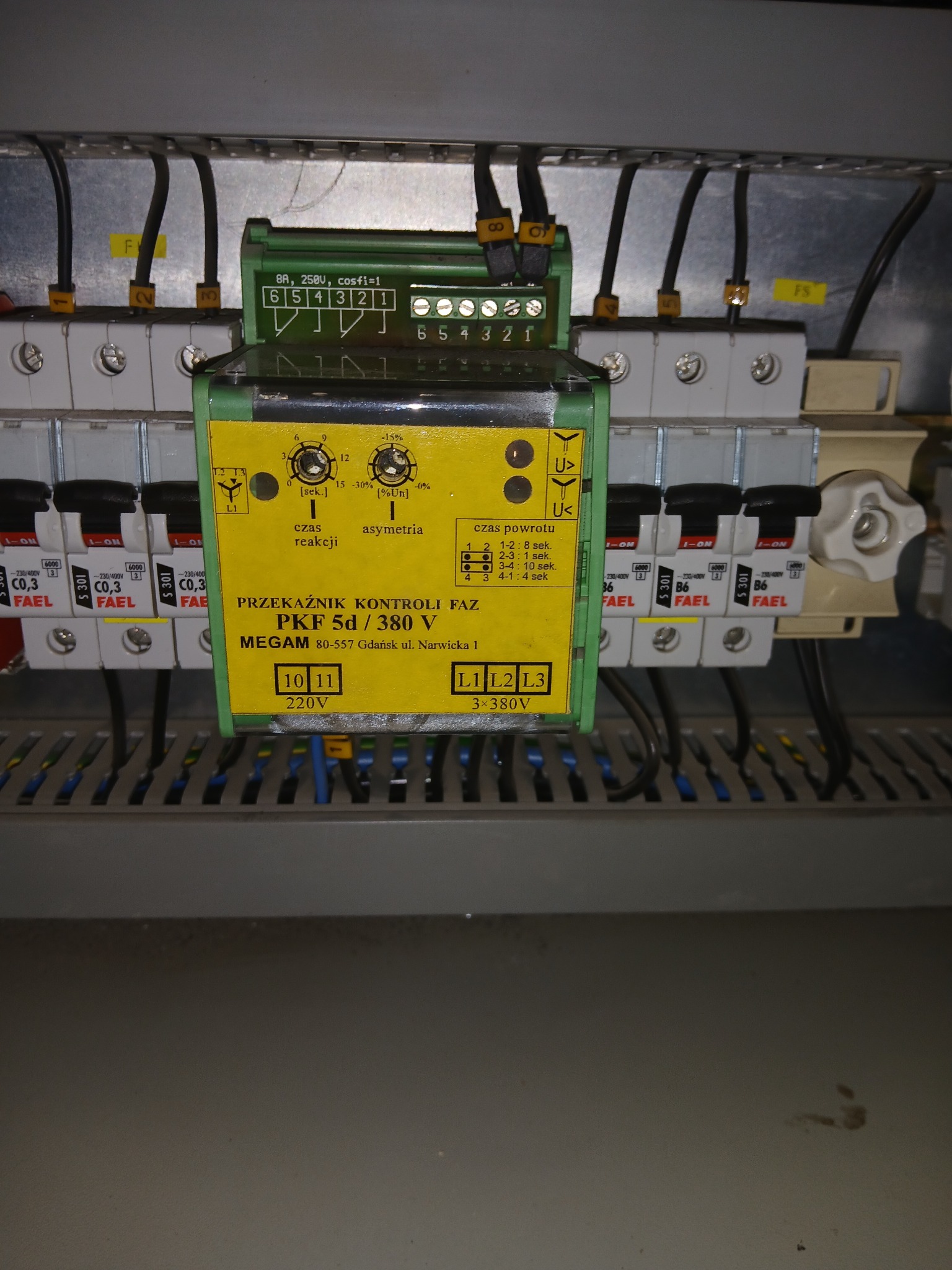 Szczegółowe ujęcie tablicy elektrycznej z przekaźnikiem kontroli faz, widoczne bezpieczniki i okablowanie. Element instalacji elektrycznej w szarej obudowie.