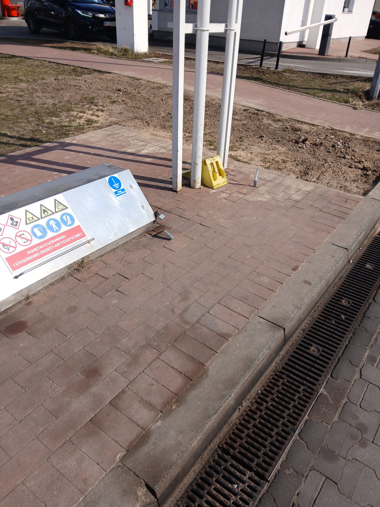 Metalowa obudowa z piktogramami BHP i napisem o odzieży antystatycznej, stojąca na czerwonej kostce brukowej obok kratki ściekowej i białych słupków.