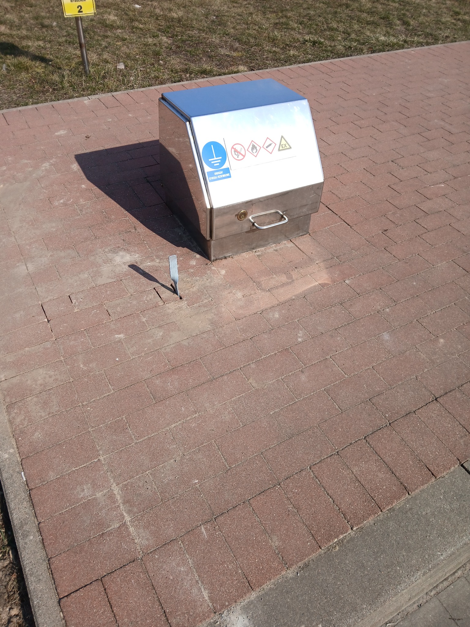 Metalowa szafka z oznaczeniami ostrzegawczymi na kostce brukowej, obok wbity metalowy pręt. Widoczny znak ostrzegający o uziemieniu. Słoneczny dzień.