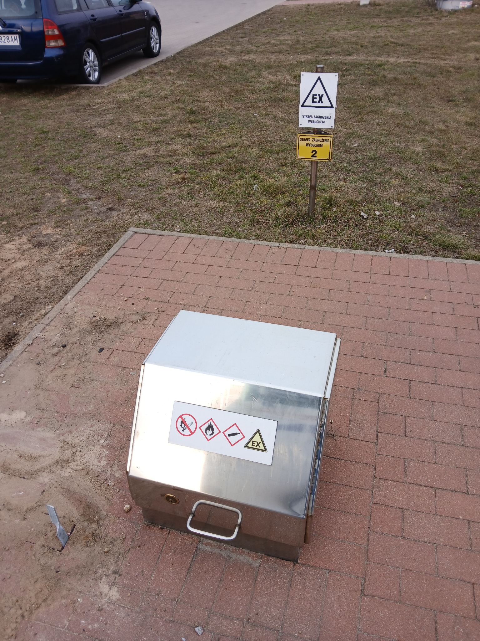 Metalowa skrzynka z oznaczeniami strefy zagrożenia wybuchem na chodniku z kostki, obok znak ostrzegawczy i trawnik. Widoczny fragment samochodu.