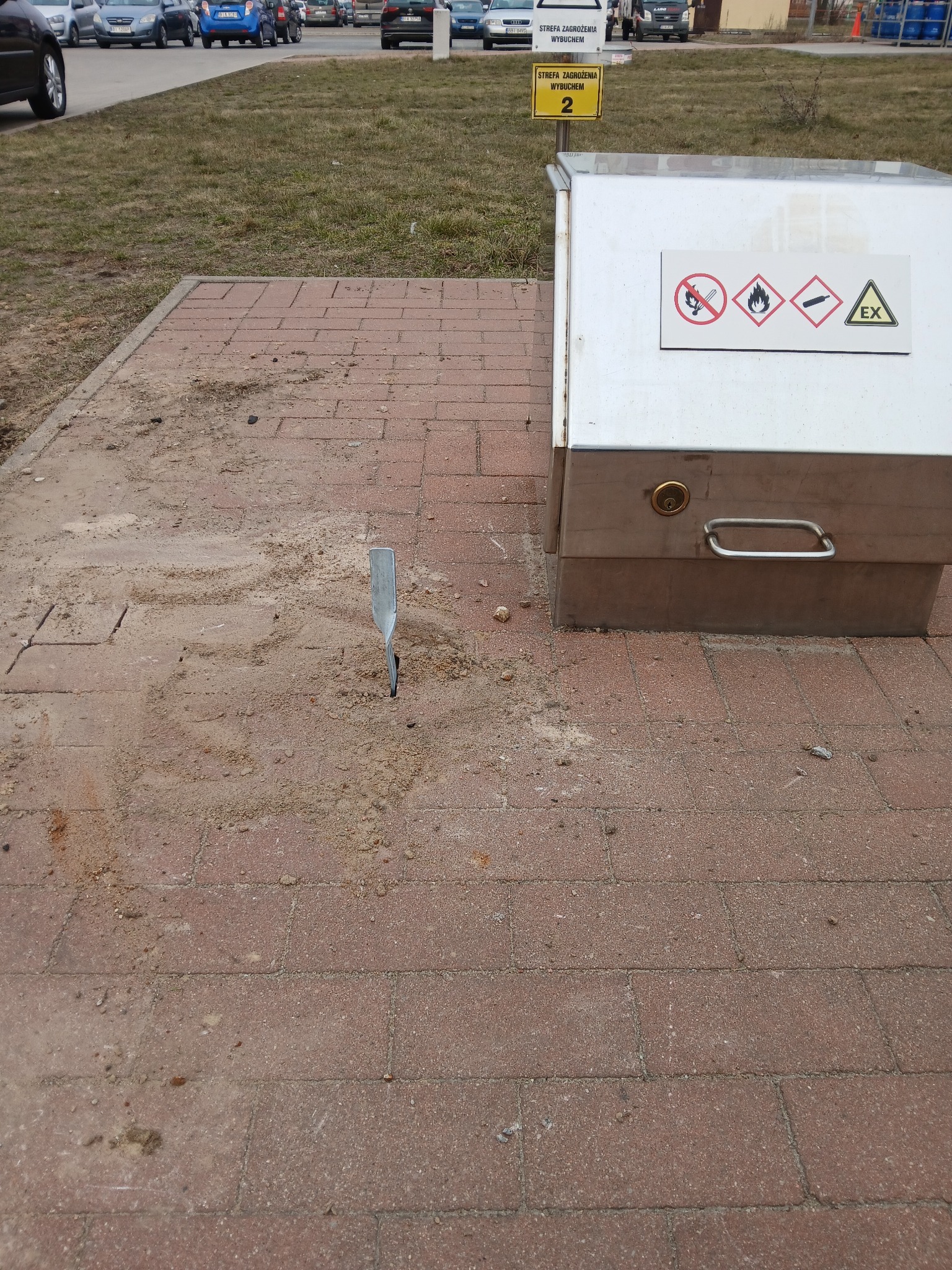 Uziemienie wbite w kostkę brukową obok metalowej szafki z oznaczeniami strefy zagrożenia wybuchem. W tle parking i budynek.