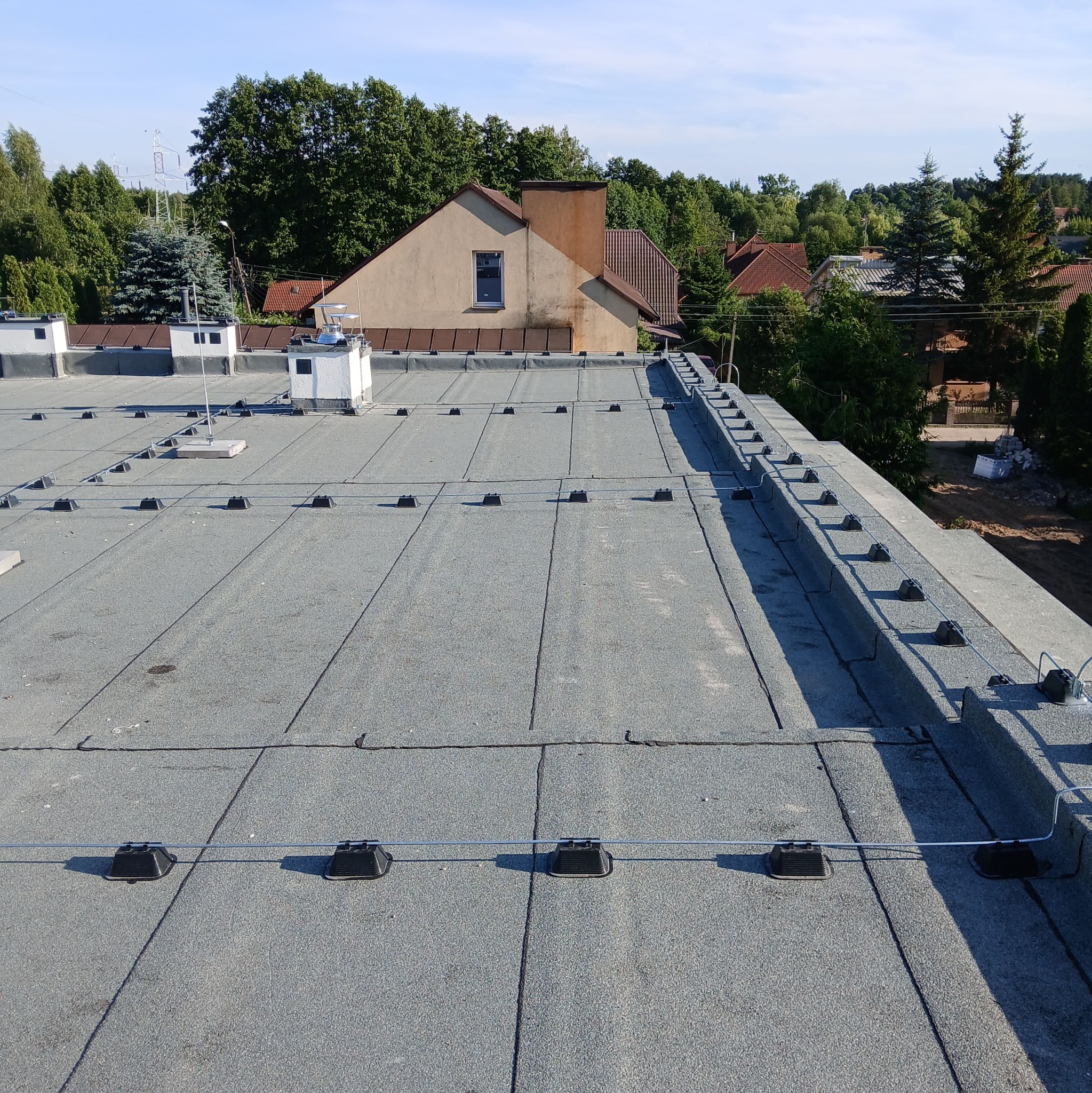 Szary dach pokryty papą z zamontowaną instalacją odgromową. Widoczne czarne uchwyty i przewody. W tle budynki i drzewa.