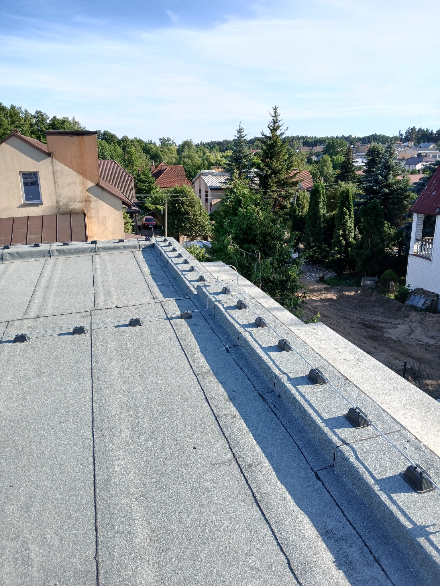 Szary dach budynku z zamontowaną instalacją odgromową, widok z góry na tle zabudowy jednorodzinnej i zieleni w słoneczny dzień. Elementy instalacji wzdłuż krawędzi dachu.