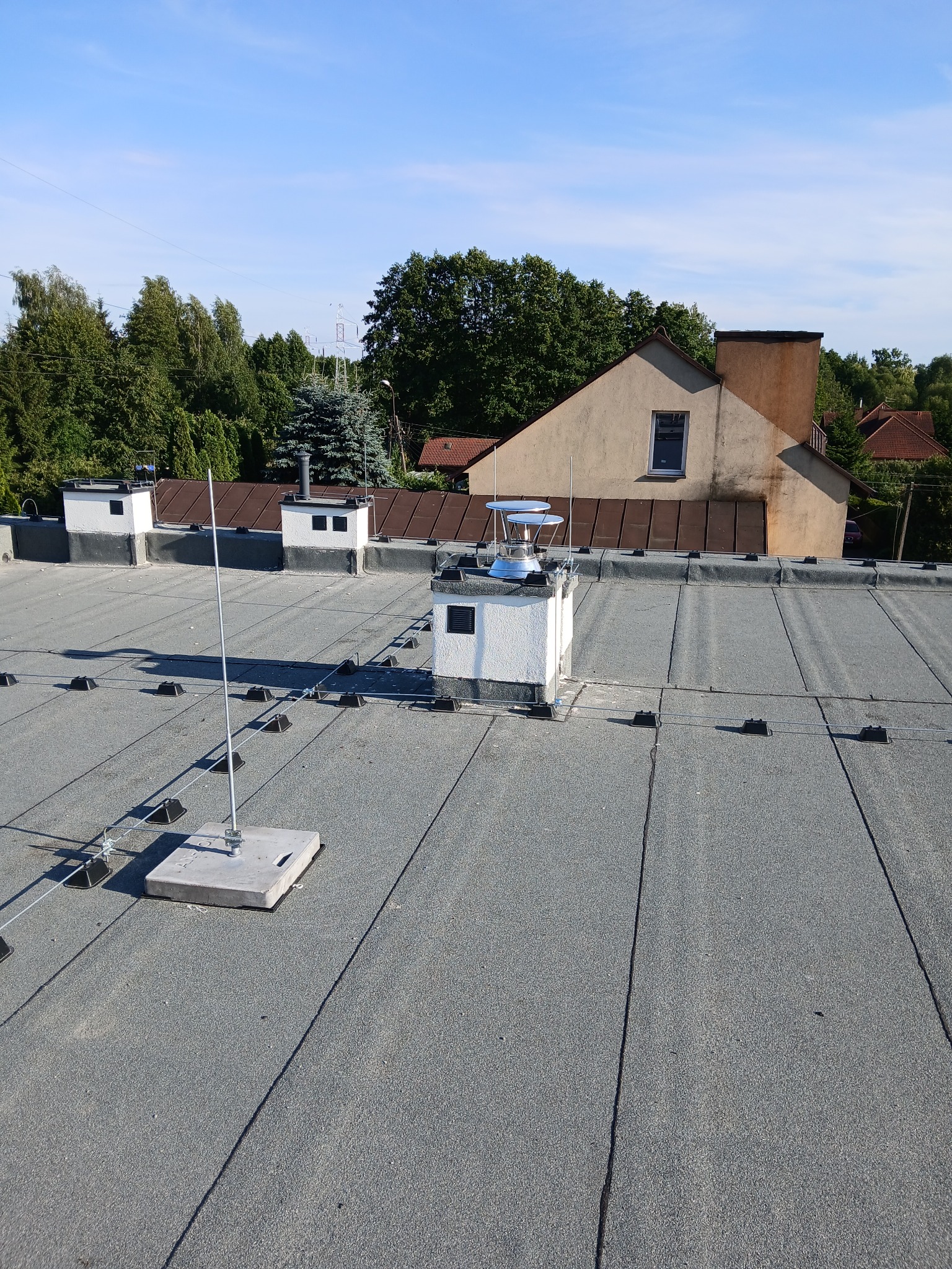 Szary dach z widoczną instalacją odgromową, z przewodami i masztami. W tle budynek i drzewa pod błękitnym niebem.