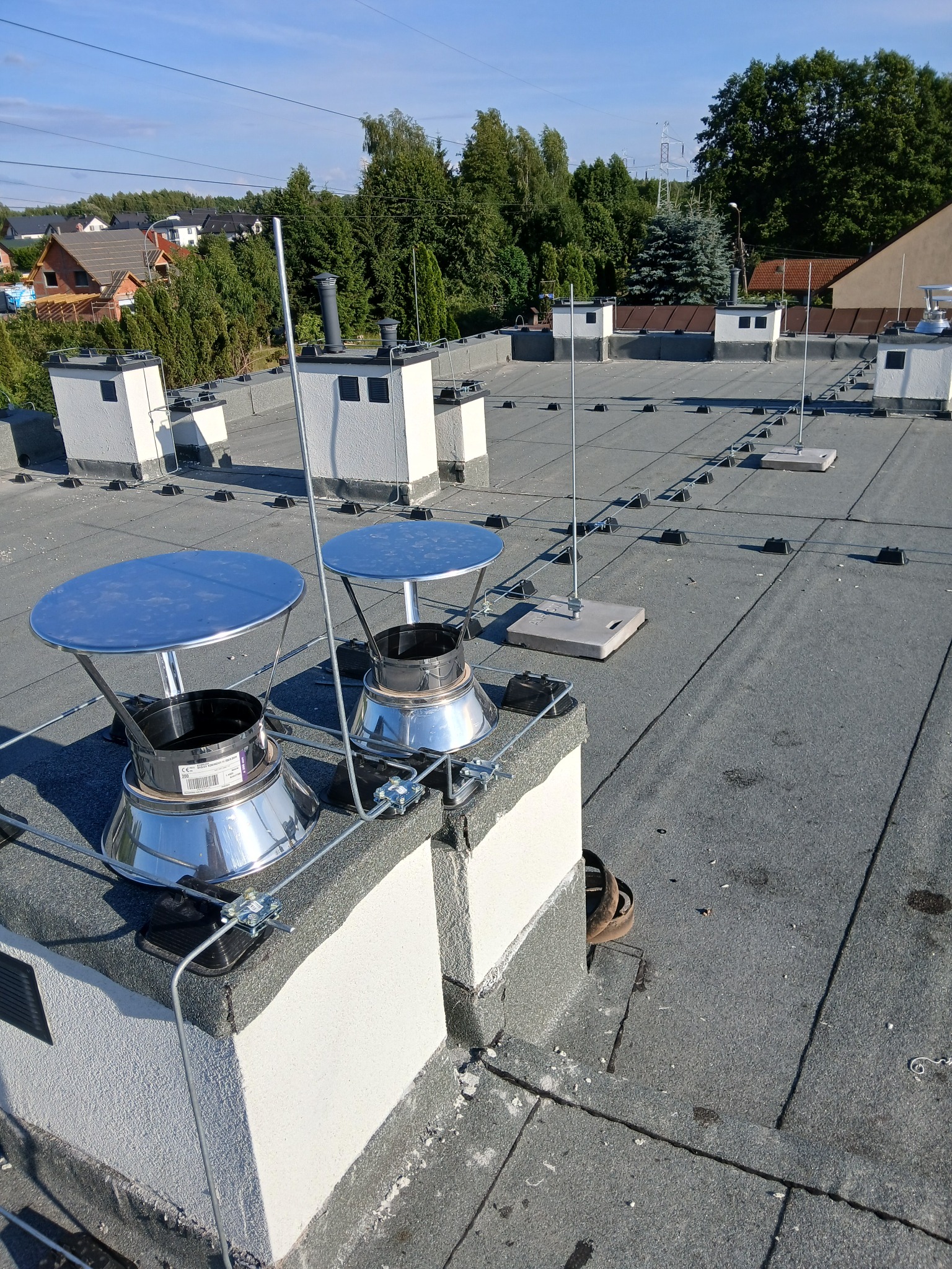 Szary dach z zainstalowaną siatką odgromową i kominami z metalowymi nasadami. Widoczne elementy mocujące i przewody. W tle zabudowa jednorodzinna i zieleń.