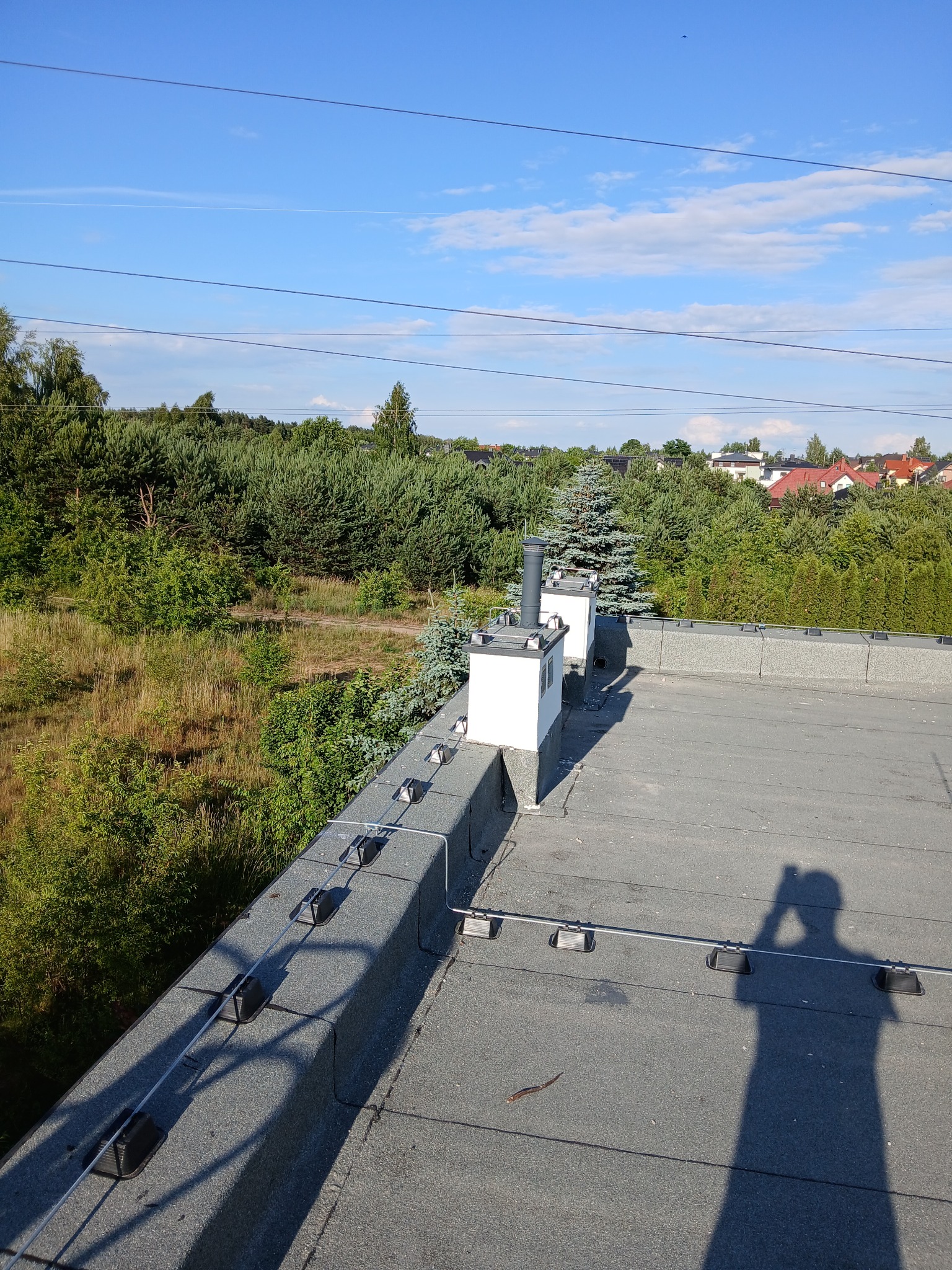 Szary dach z widoczną instalacją odgromową, tłem jest zieleń drzew i zabudowa mieszkalna. Na dachu cień osoby robiącej zdjęcie.