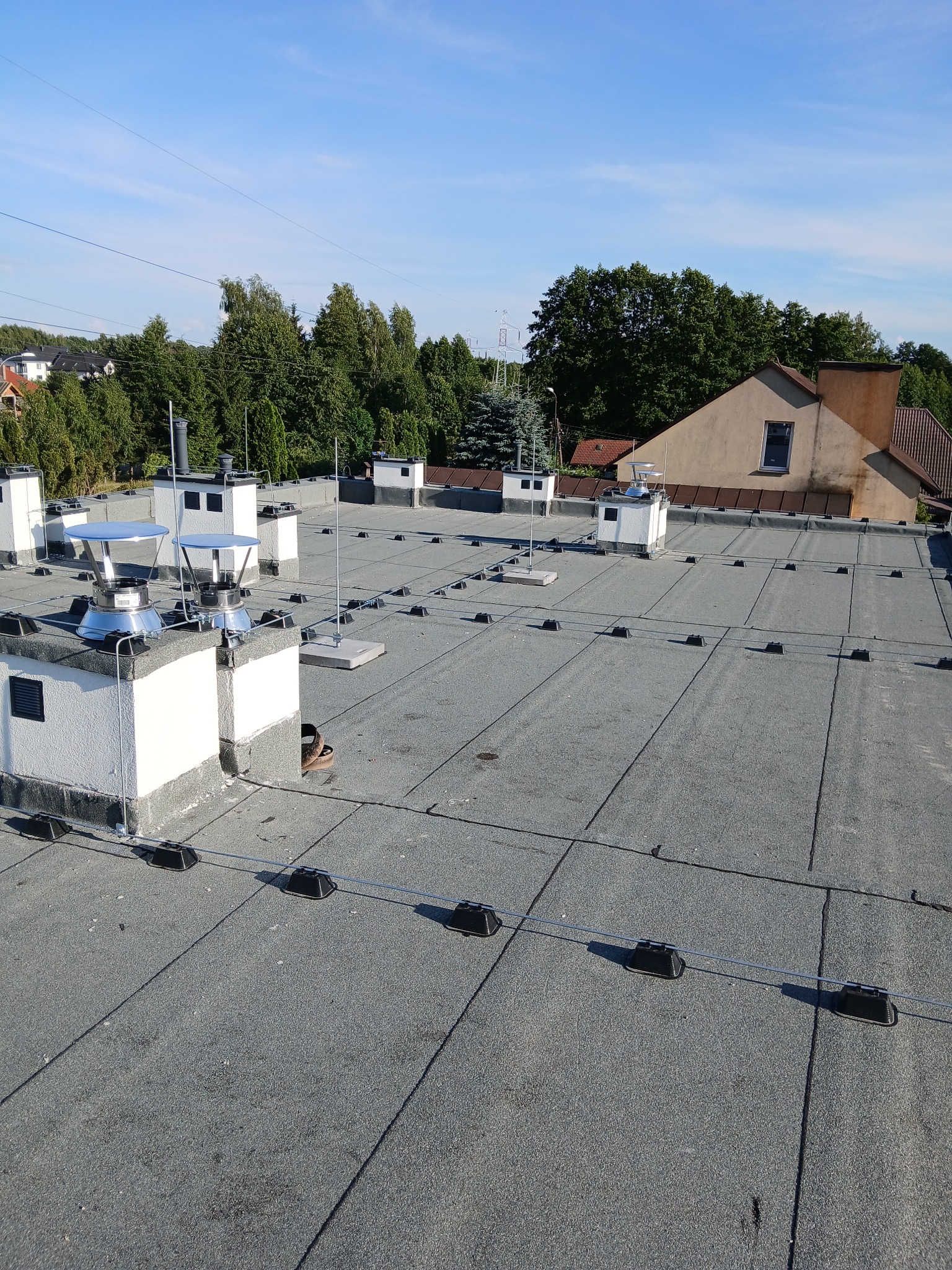 Szary dach pokryty papą z zamontowaną instalacją odgromową, widoczne kominy i przewody odgromowe na tle zieleni i błękitnego nieba.