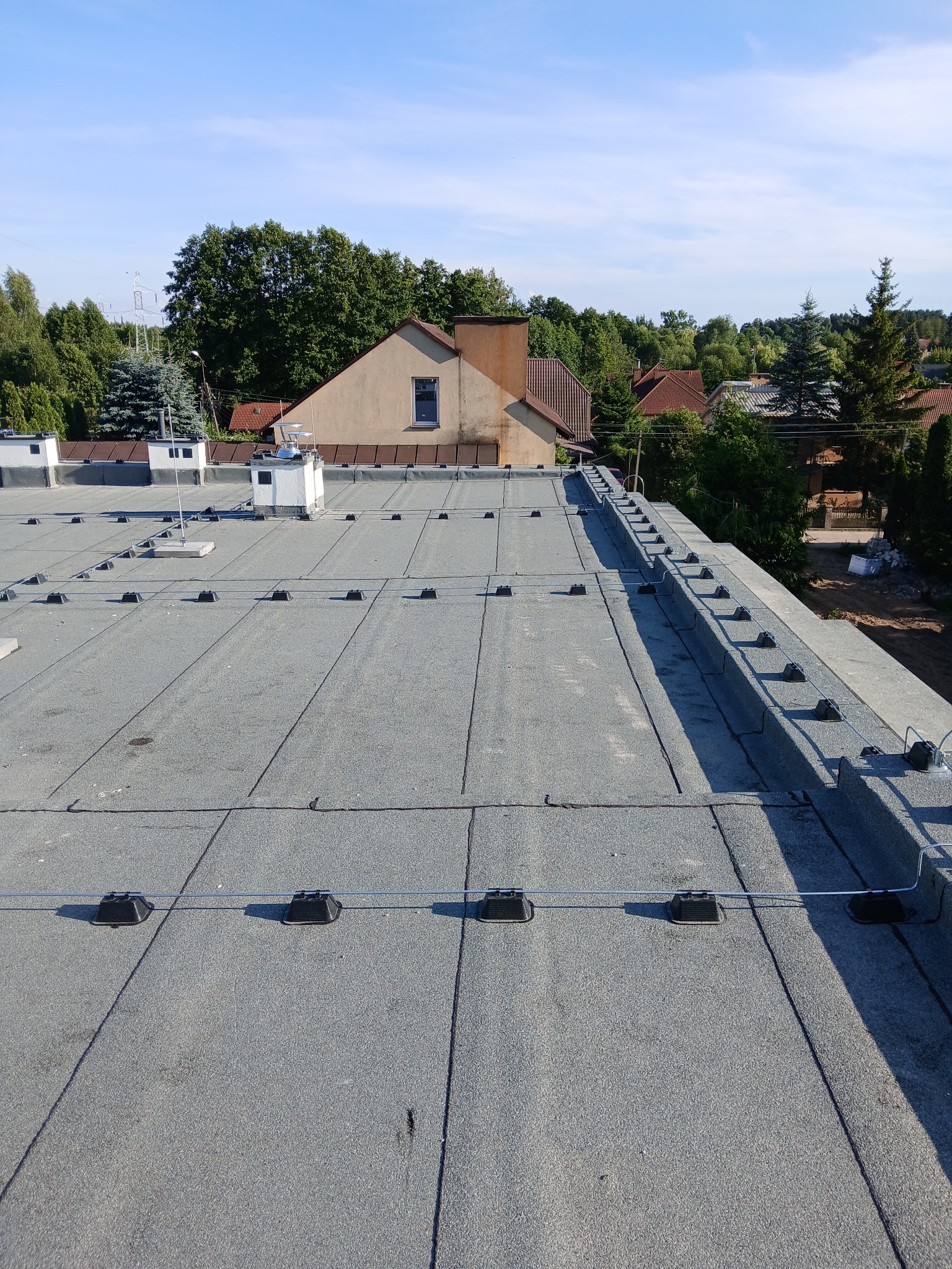 Szary dach budynku z zamontowaną instalacją odgromową, widoczne przewody i uchwyty. W tle zabudowa jednorodzinna i zieleń.