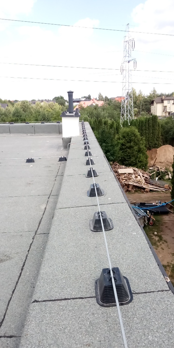 Szary dach z zamontowaną instalacją odgromową, widoczne uchwyty i przewód. W tle komin, drzewa i słup wysokiego napięcia.