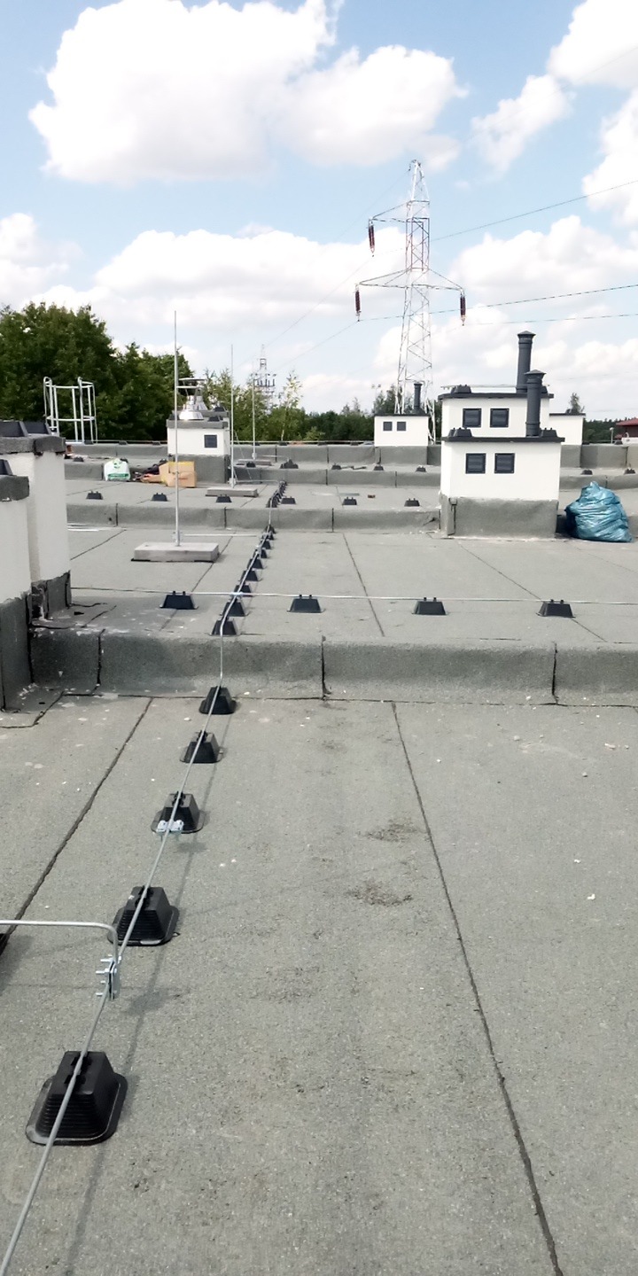 Dach pokryty papą z zamontowaną instalacją odgromową, widoczne przewody i uchwyty. W tle słupy wysokiego napięcia, Białystok.