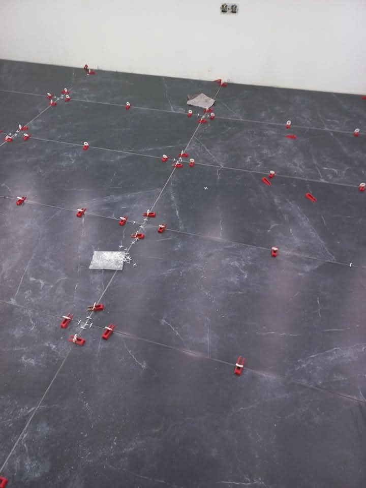 Układanie ciemnych płytek podłogowych z użyciem czerwonych klipsów poziomujących. Widoczne fugi i fragment ściany z gniazdkami elektrycznymi.