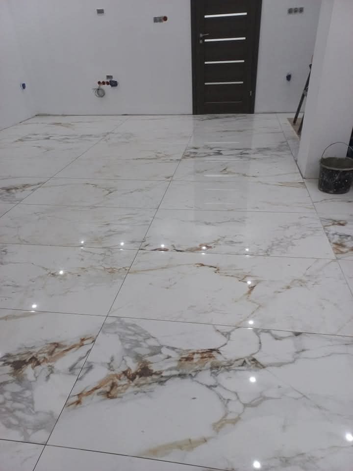 Położona podłoga z płytek imitujących marmur w jasnym pomieszczeniu z ciemnymi drzwiami. Widoczne fugi i odbicia światła na powierzchni płytek.