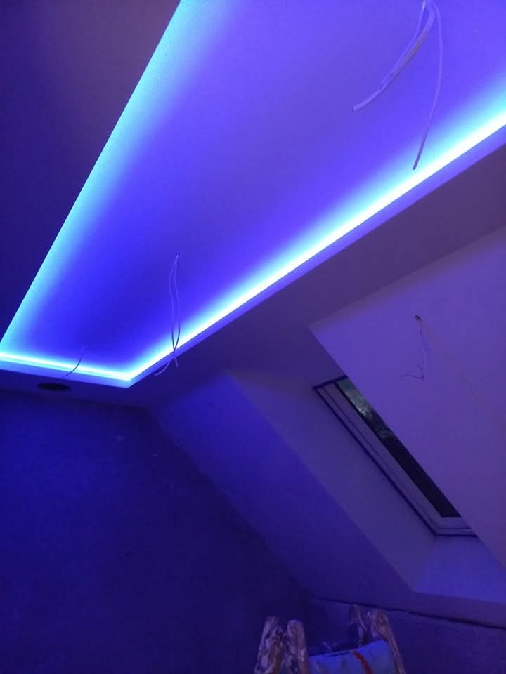 Sufit podwieszany z oświetleniem LED w kolorze niebieskim, widok z dołu, fragment okna dachowego i drabiny malarskiej w rogu kadru. Nowoczesne wykończenie.