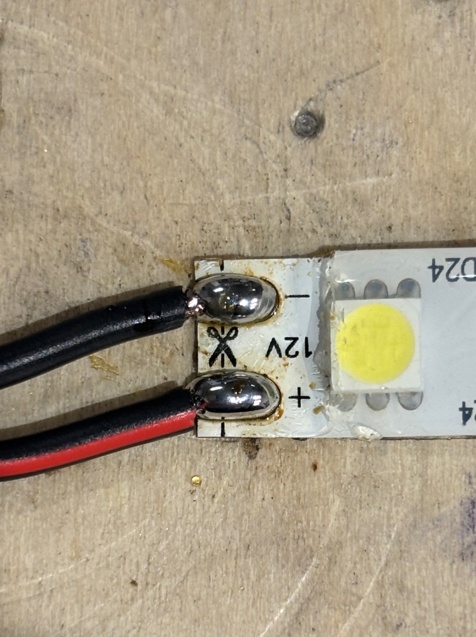 Zbliżenie na diodę LED z przylutowanymi przewodami (czerwony i czarny) na drewnianym podłożu. Widoczne oznaczenia napięcia 12V i symbol nożyczek.