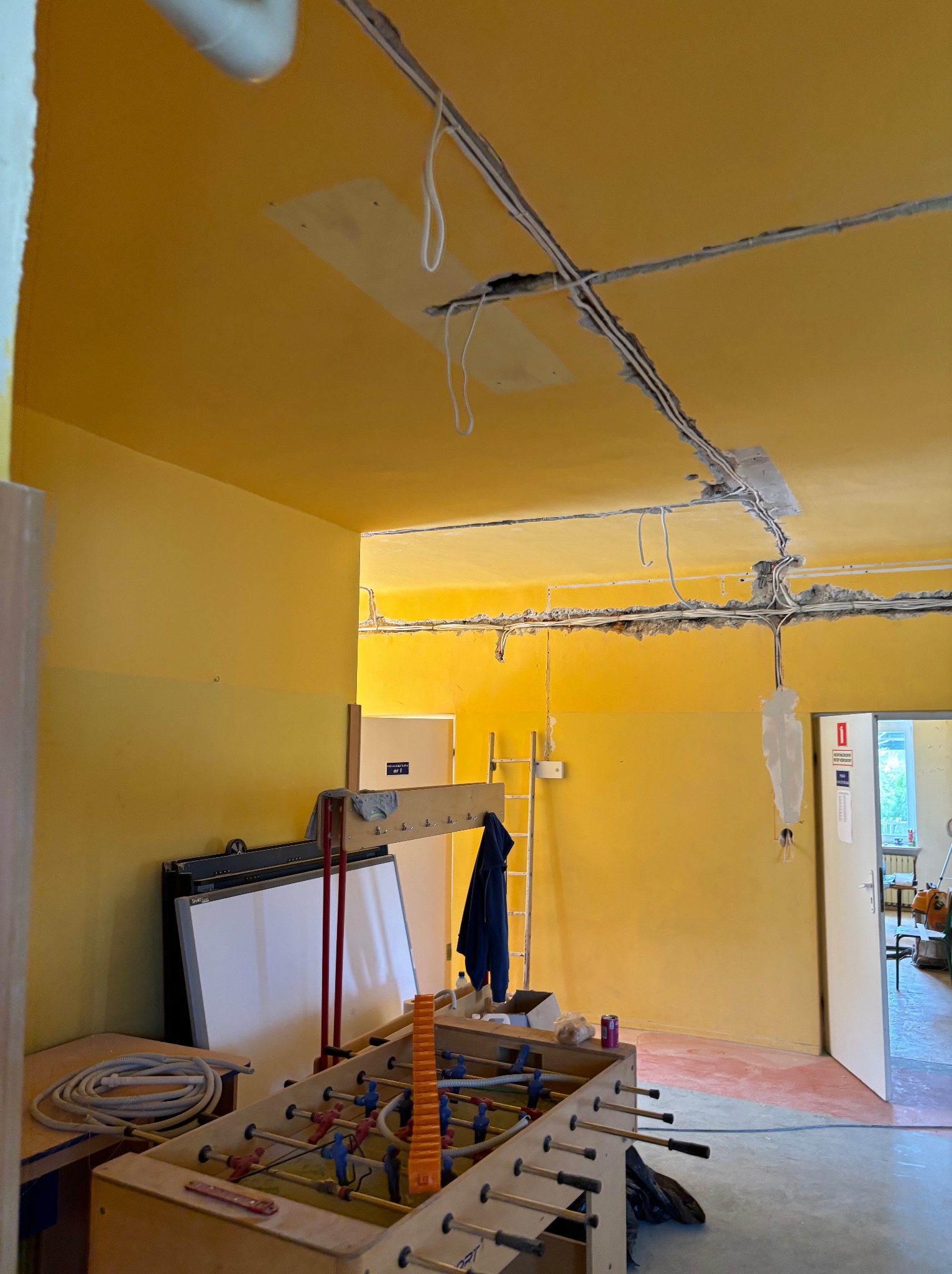 Żółte wnętrze w trakcie prac instalacyjnych: widoczne kable elektryczne w bruzdach na suficie, stół do piłkarzyków i drabina. Etap przed wykończeniem.