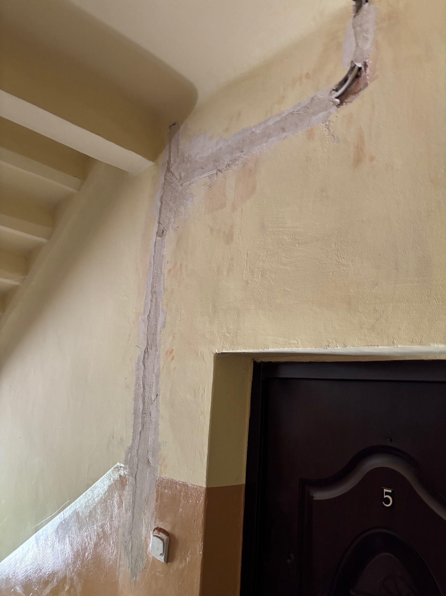 Ściana klatki schodowej z poprowadzonymi przewodami elektrycznymi w korytku i zamaskowanymi gipsem. Widoczny fragment drzwi z numerem mieszkania.