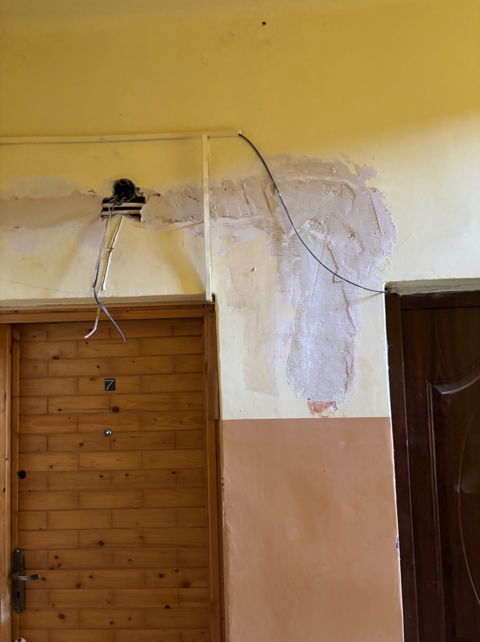 Otwór w ścianie z wystającymi kablami, obok drewniane drzwi z numerem 7. Ściana częściowo pokryta gładzią, widoczne ślady po instalacji elektrycznej.