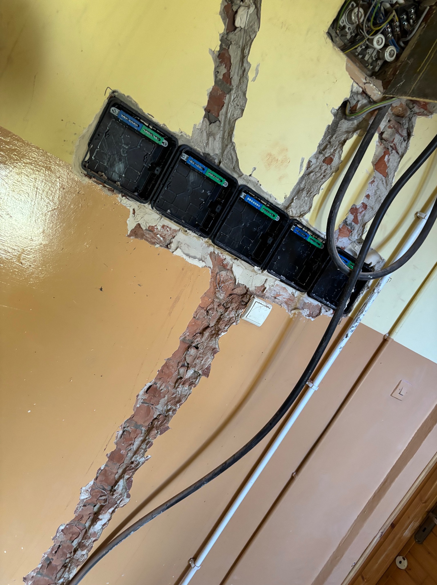 Rząd czarnych puszek instalacyjnych w ścianie z odsłoniętą cegłą, widoczne kable i przewody elektryczne. Żółto-brązowa ściana w tle, ujęcie pod kątem.