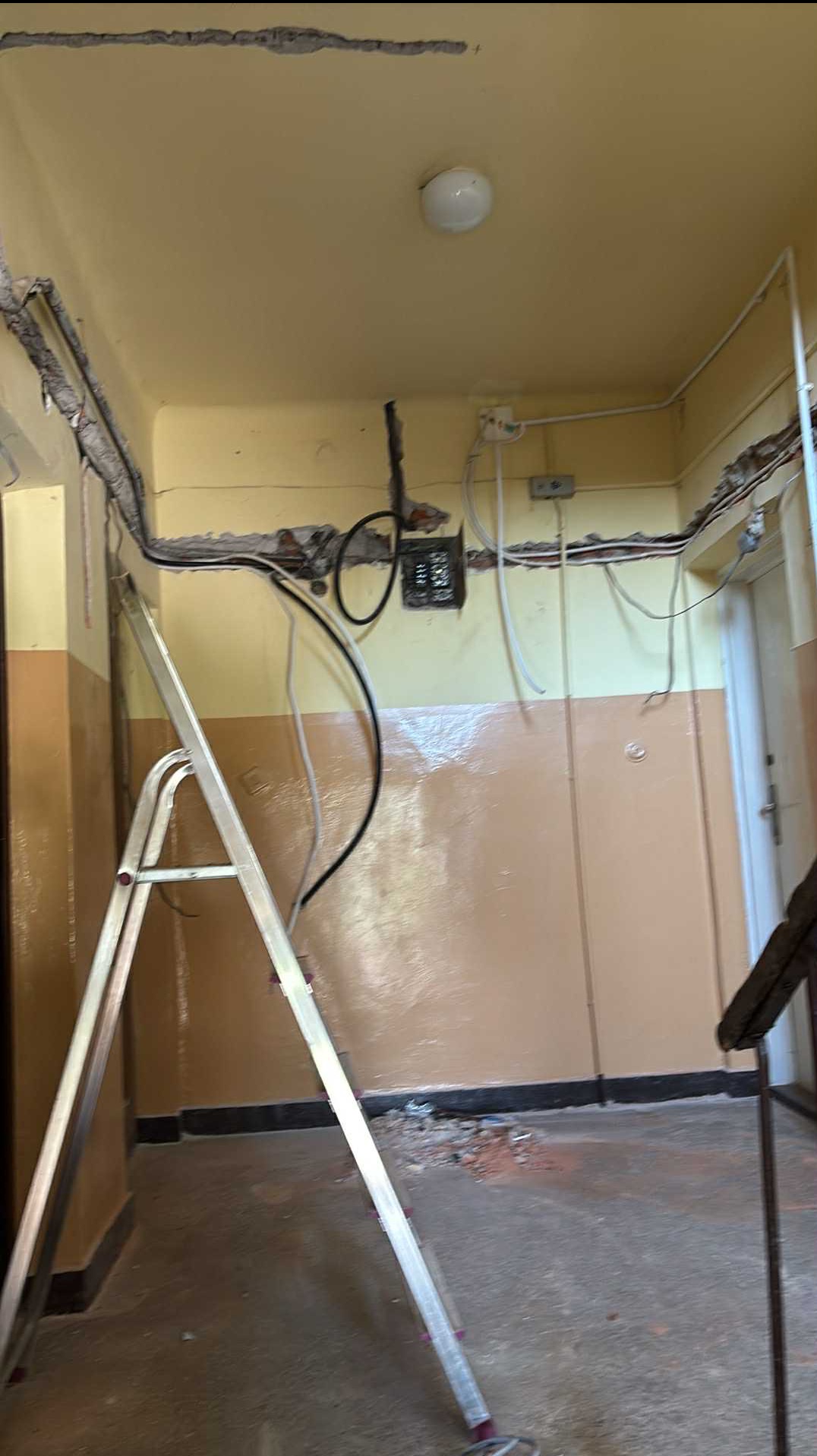 Korytarz w trakcie remontu instalacji elektrycznej: widoczne kable, puszki elektryczne, drabina aluminiowa. Ściany dwukolorowe, oświetlenie sufitowe.